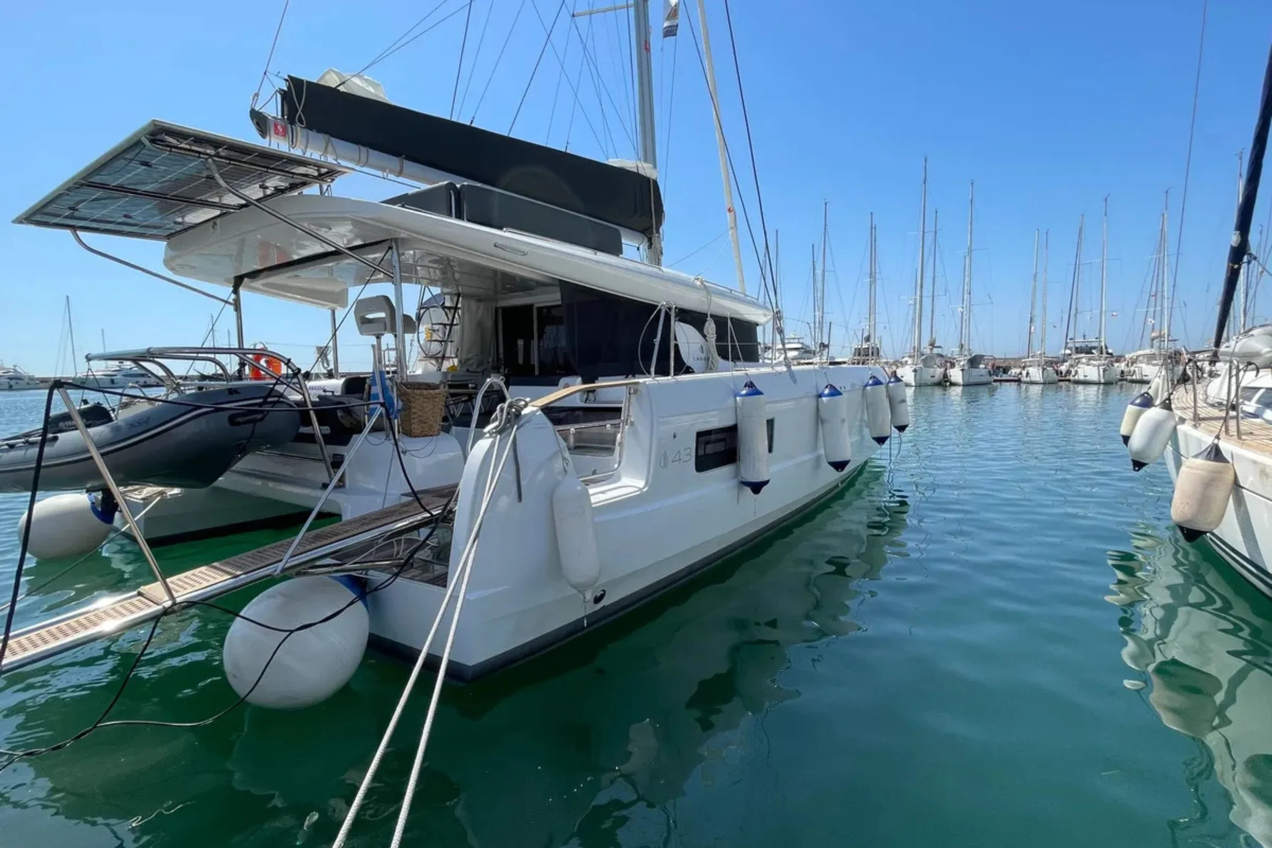 A white luxury catamaran docked in a marina with several sailboats in the background under a bright blue sky.