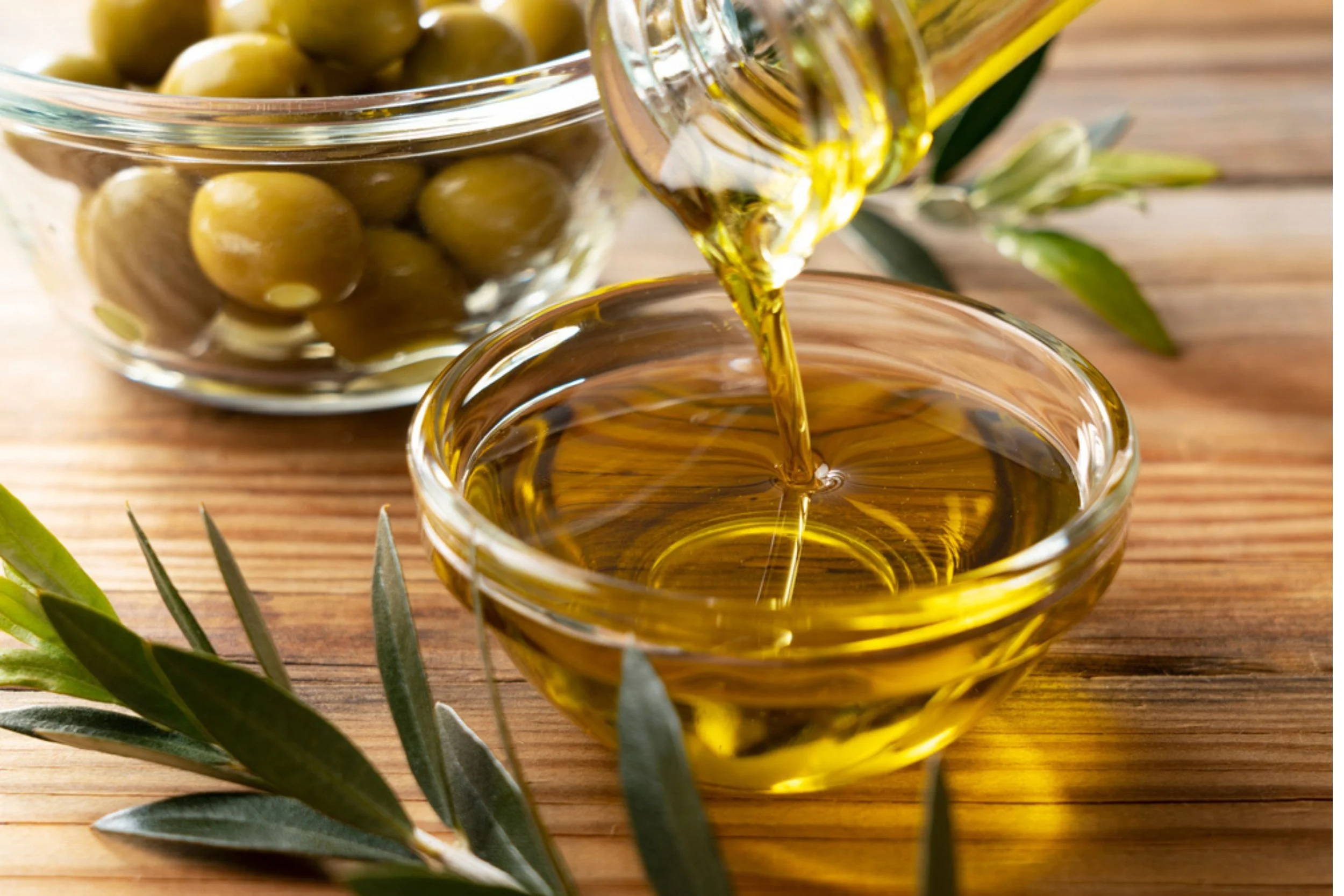 Olive oil being poured from a bottle into a glass bowl on a wooden surface, with a jar of green olives and olive leaves nearby.