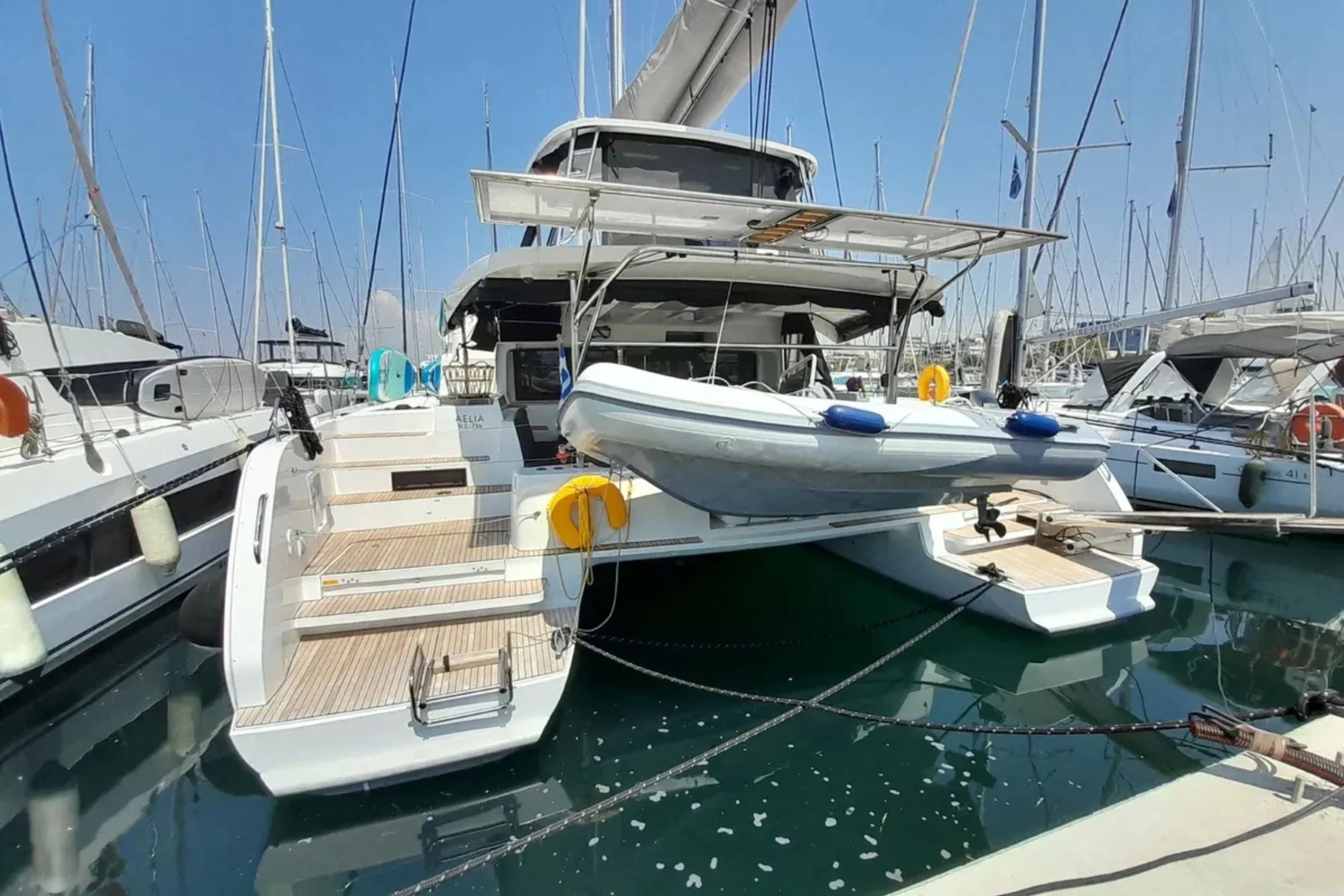 A white yacht docked at a marina, with a smaller boat hanging on its side, surrounded by other sailboats. The sky is blue with some clouds.