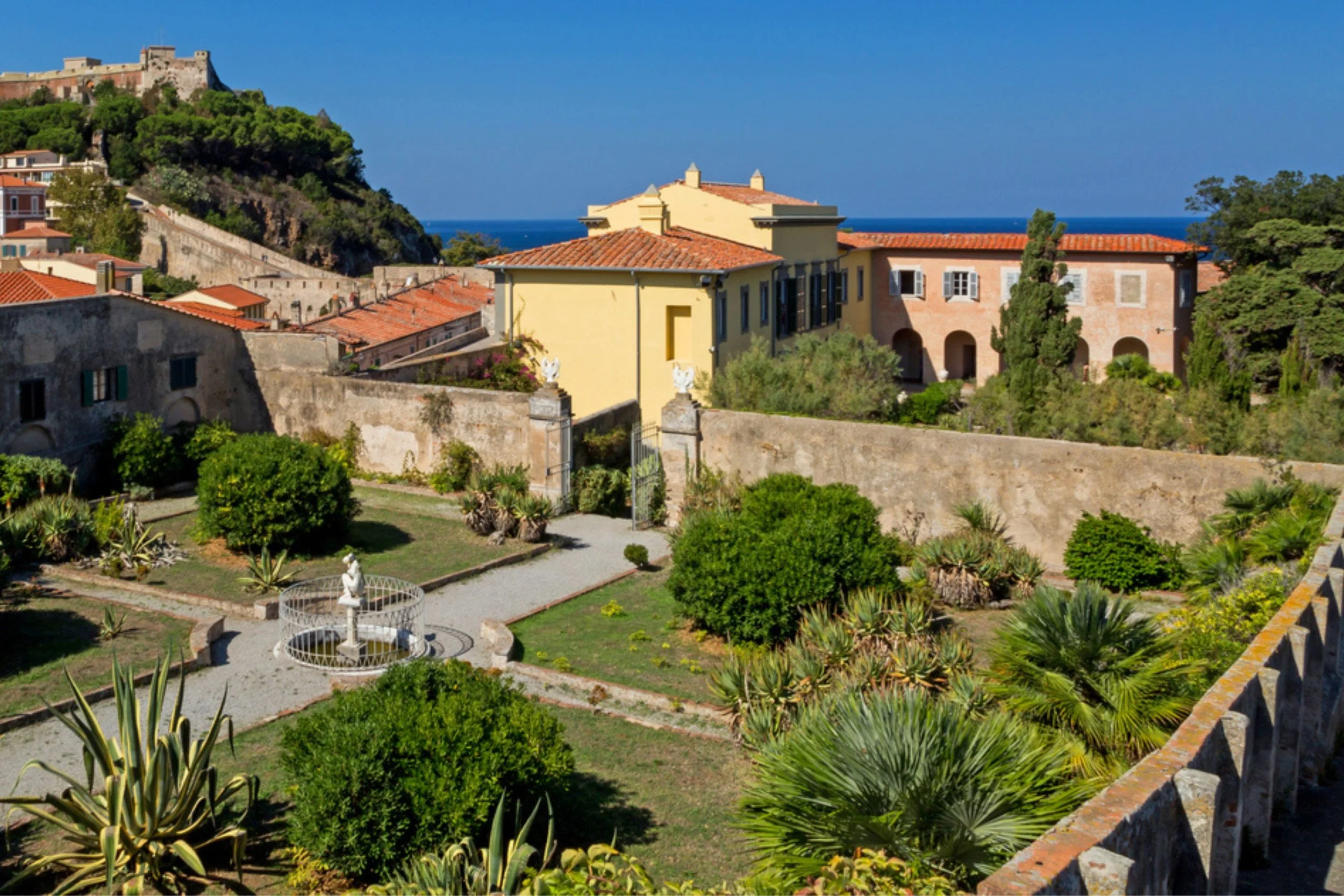 Scenic view of a lush courtyard with a statue fountain, surrounded by colorful buildings and greenery, with a distant view of the sea and hills in the background.