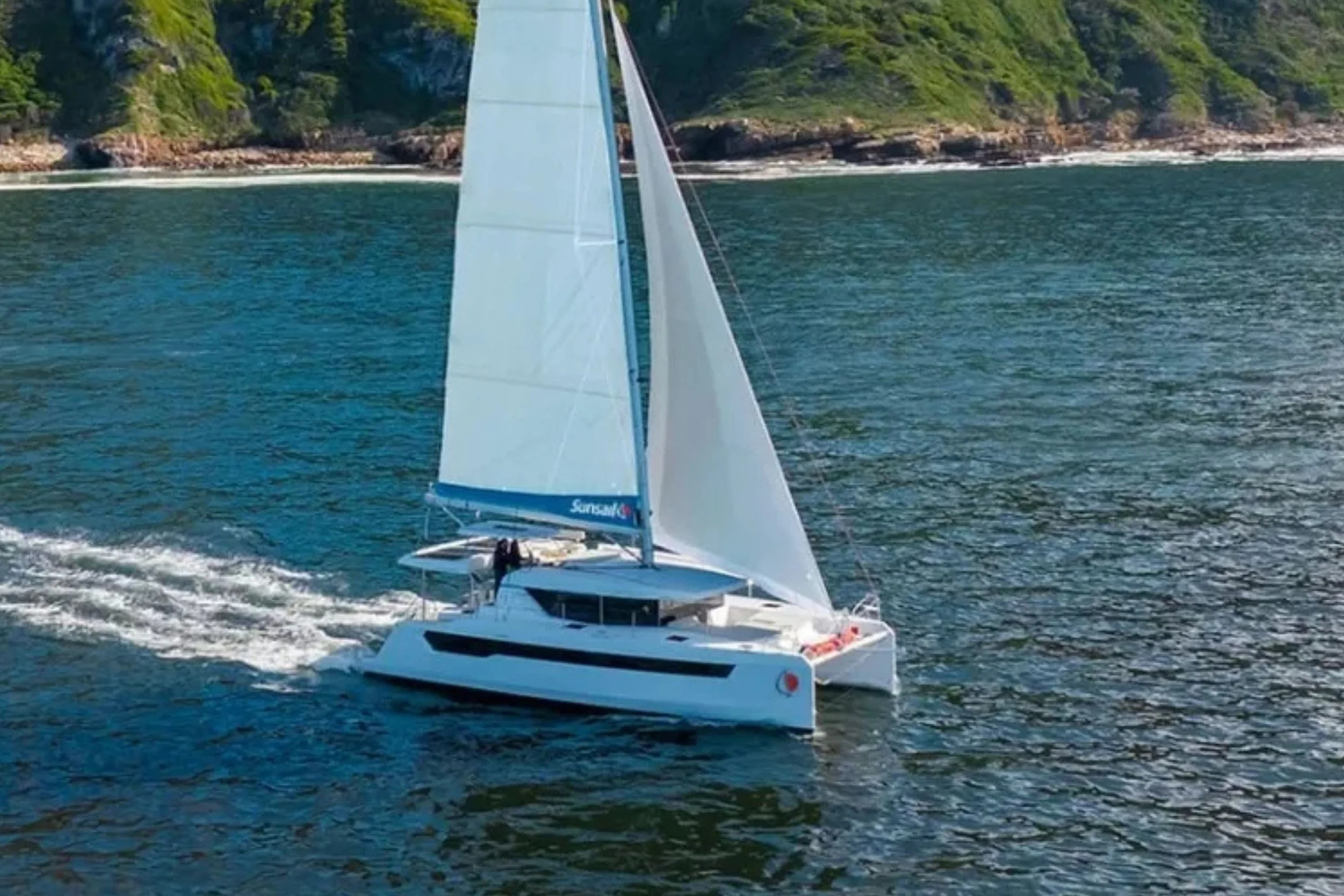 A white sailboat with its sails up is sailing on blue water near a lush, green coastline with rocky cliffs.
