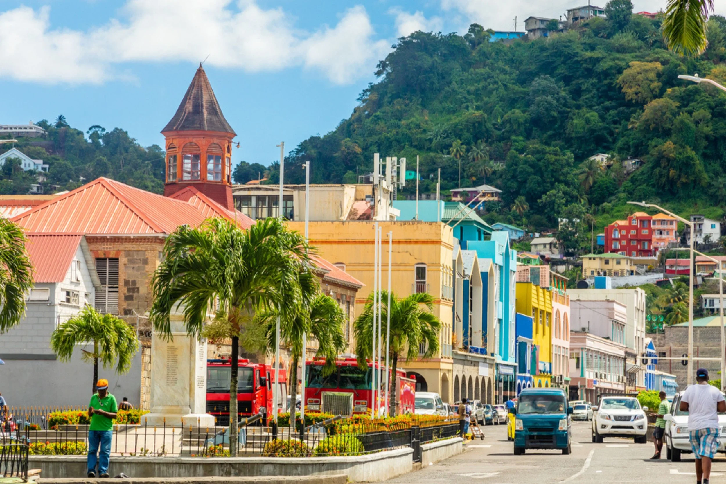 A colorful city street with buildings in shades of blue, yellow, and white, palm trees, a church with a red roof and tower, red fire trucks, cars, and people walking, with a green hill and houses in the background.