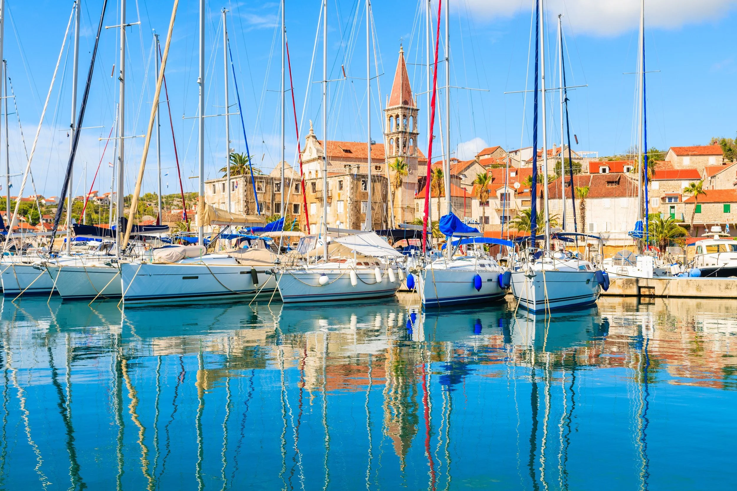 Several sailboats docked at a marina with colorful town buildings and a church with a steeple in the background, under a bright blue sky.