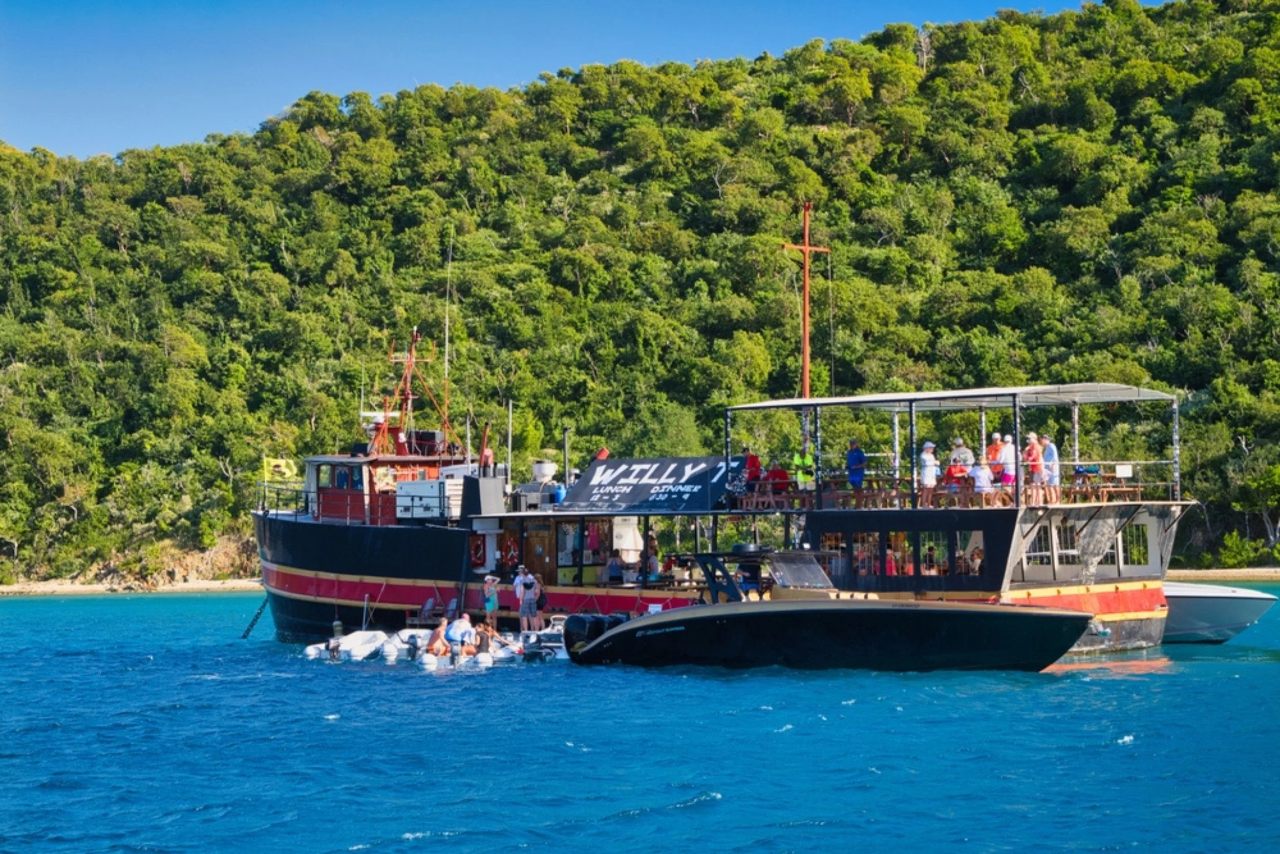 A boat named 'Willy' floating on blue water near a lush green hillside with people on board and in the water, some engaged in water activities with a black speedboat attached.