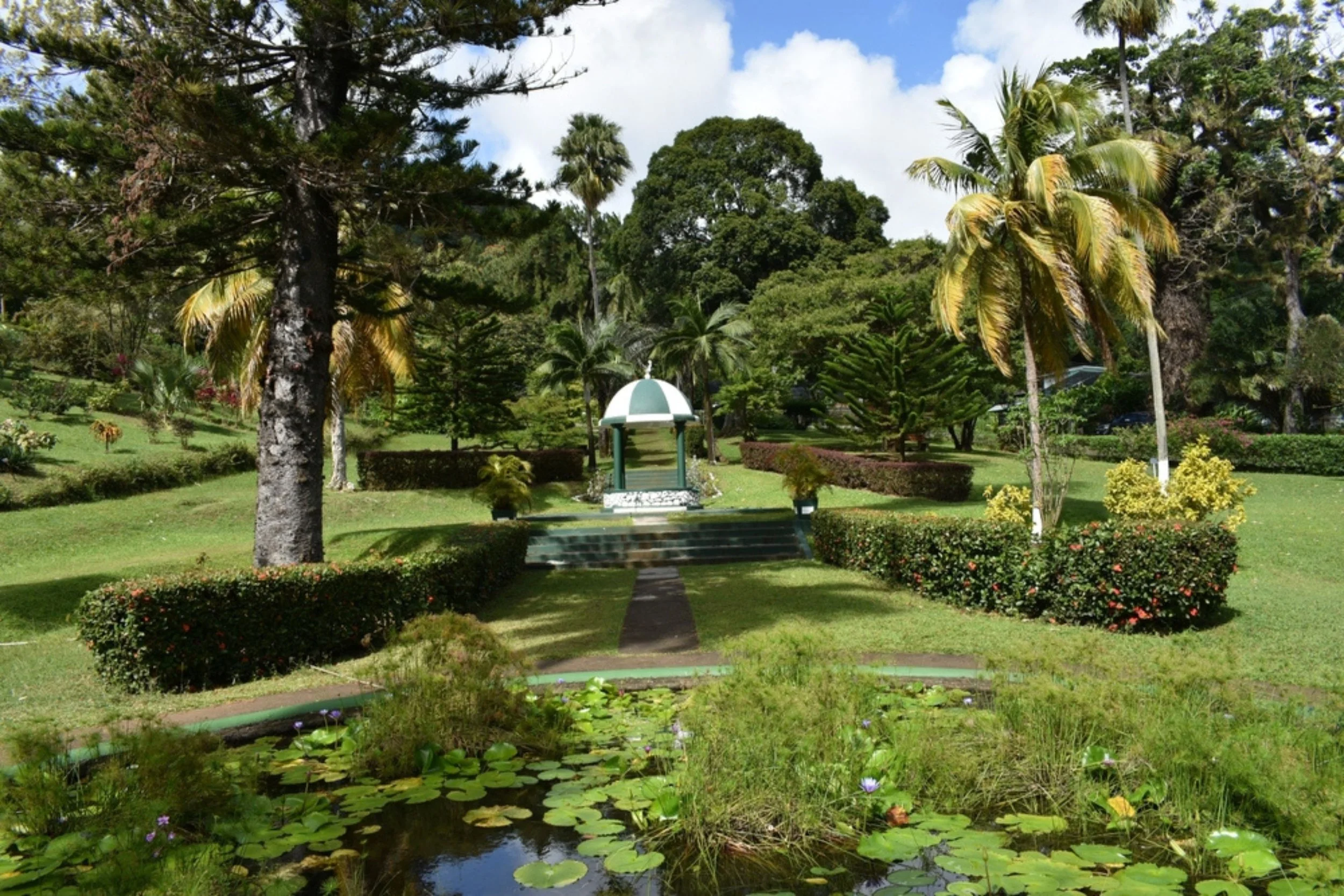 A lush garden with a pond and water lilies in the foreground. A pathway leads to a small pavilion with a domed roof, surrounded by various trees and manicured bushes.
