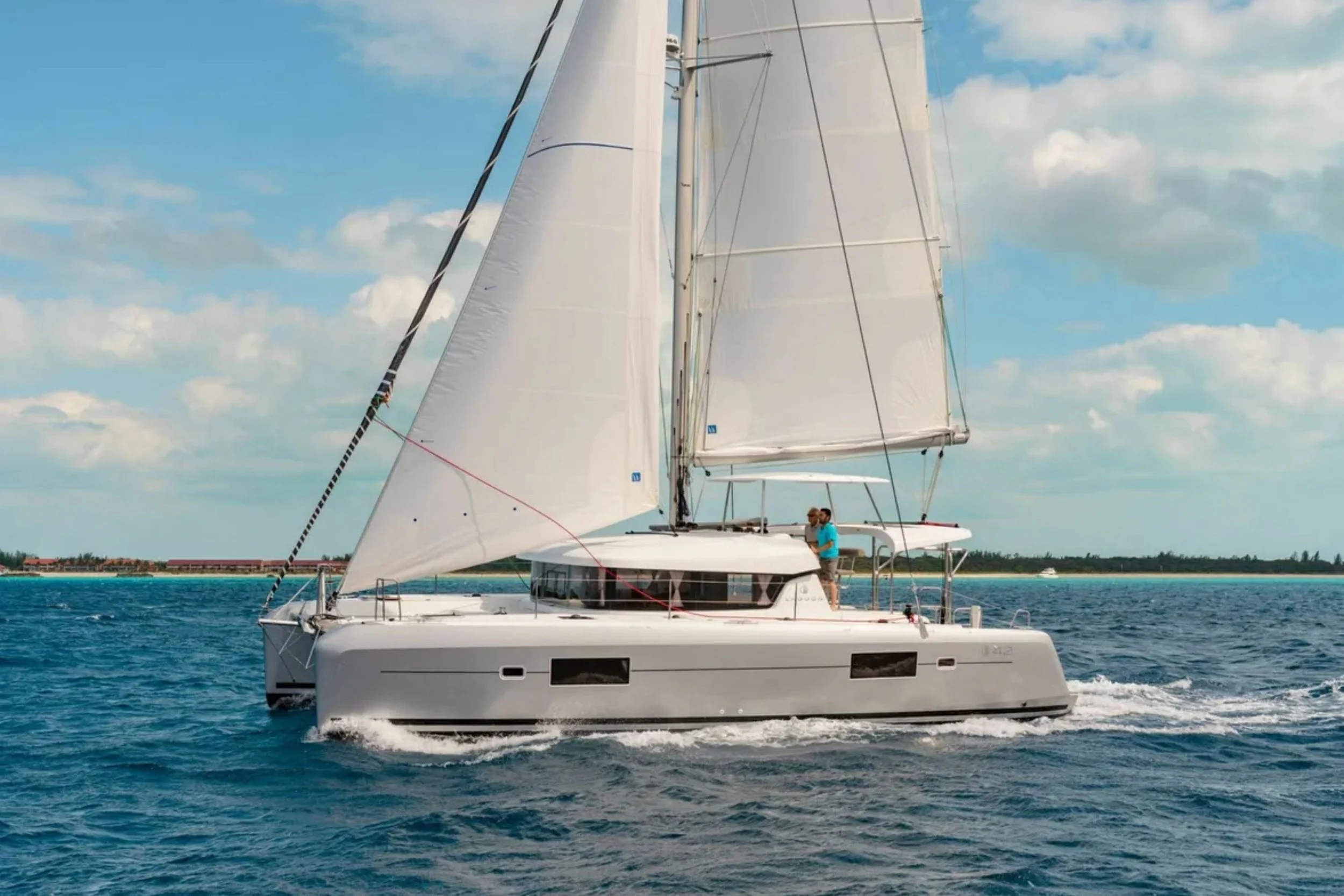 A modern white sailing catamaran with sails up, cruising on the ocean with a man on deck.