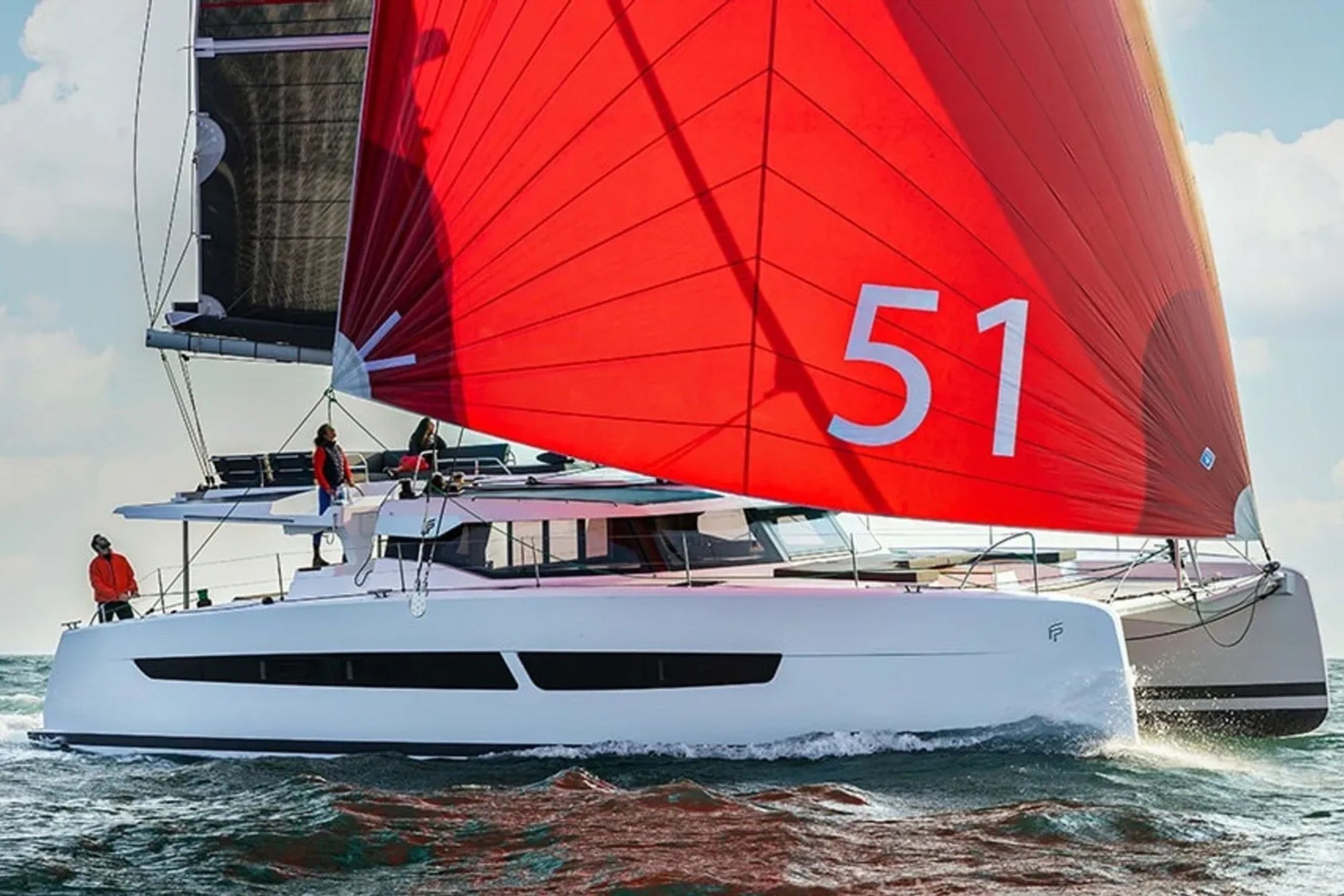 A modern sailboat with a large red sail numbered 51, sailing on the water with a partly cloudy sky in the background and four crew members on board.