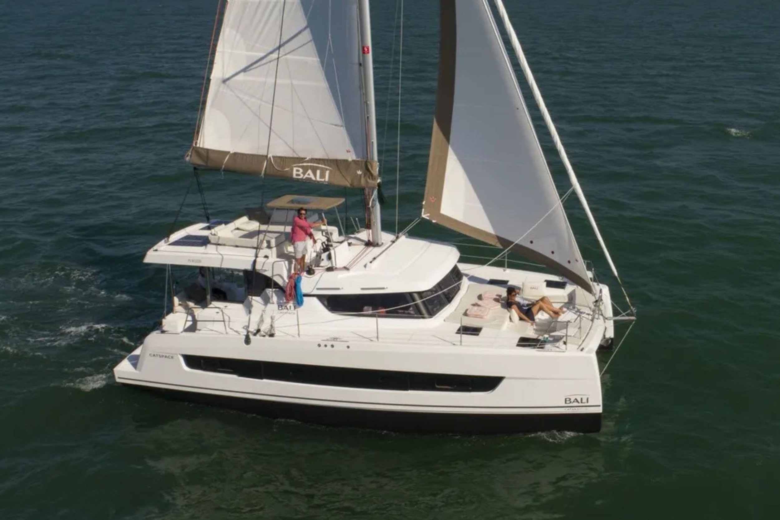 A white sailboat named Bali with two people on board sailing on the water with its sails up.