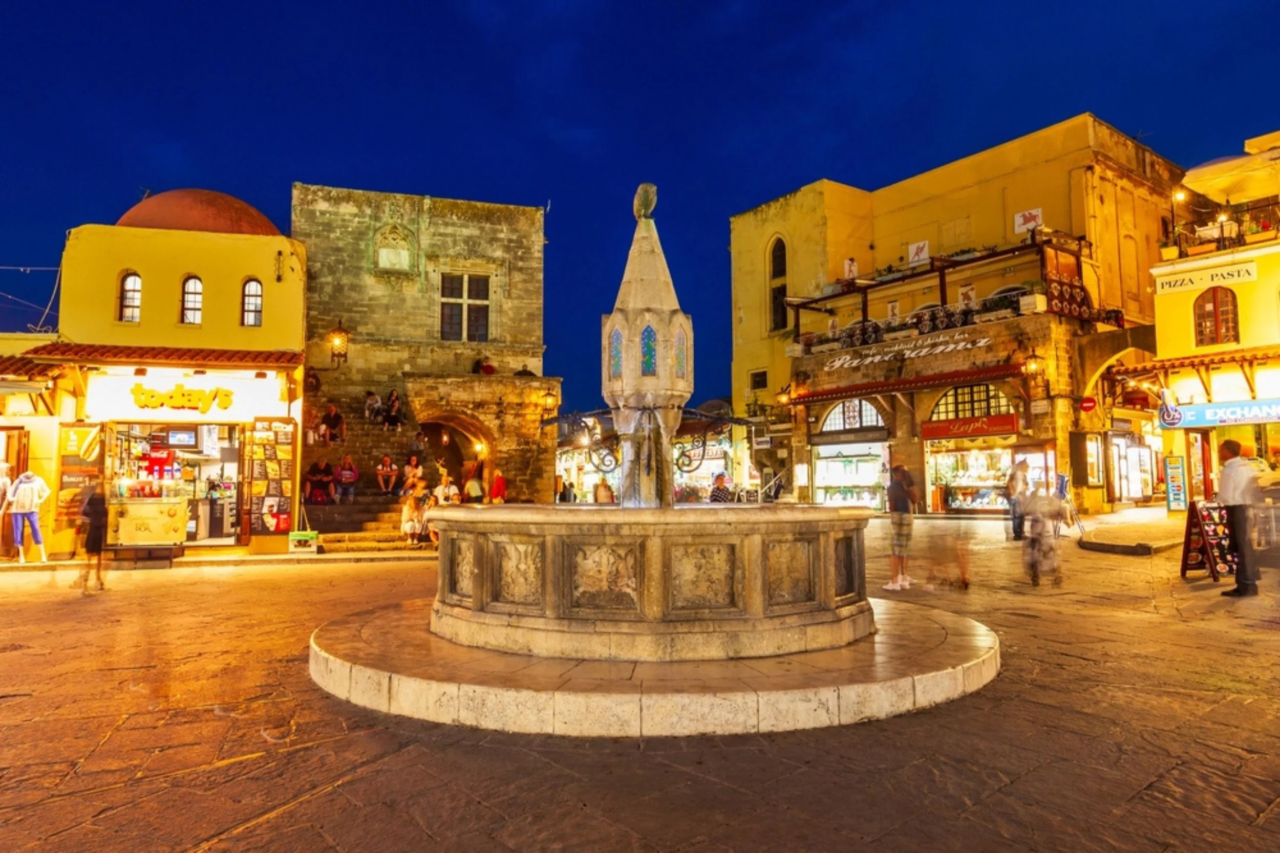 A nighttime scene of a lively city square with a stone fountain in the center, surrounded by illuminated shops, cafes, and historic buildings, with people walking and sitting on the steps near the fountain.