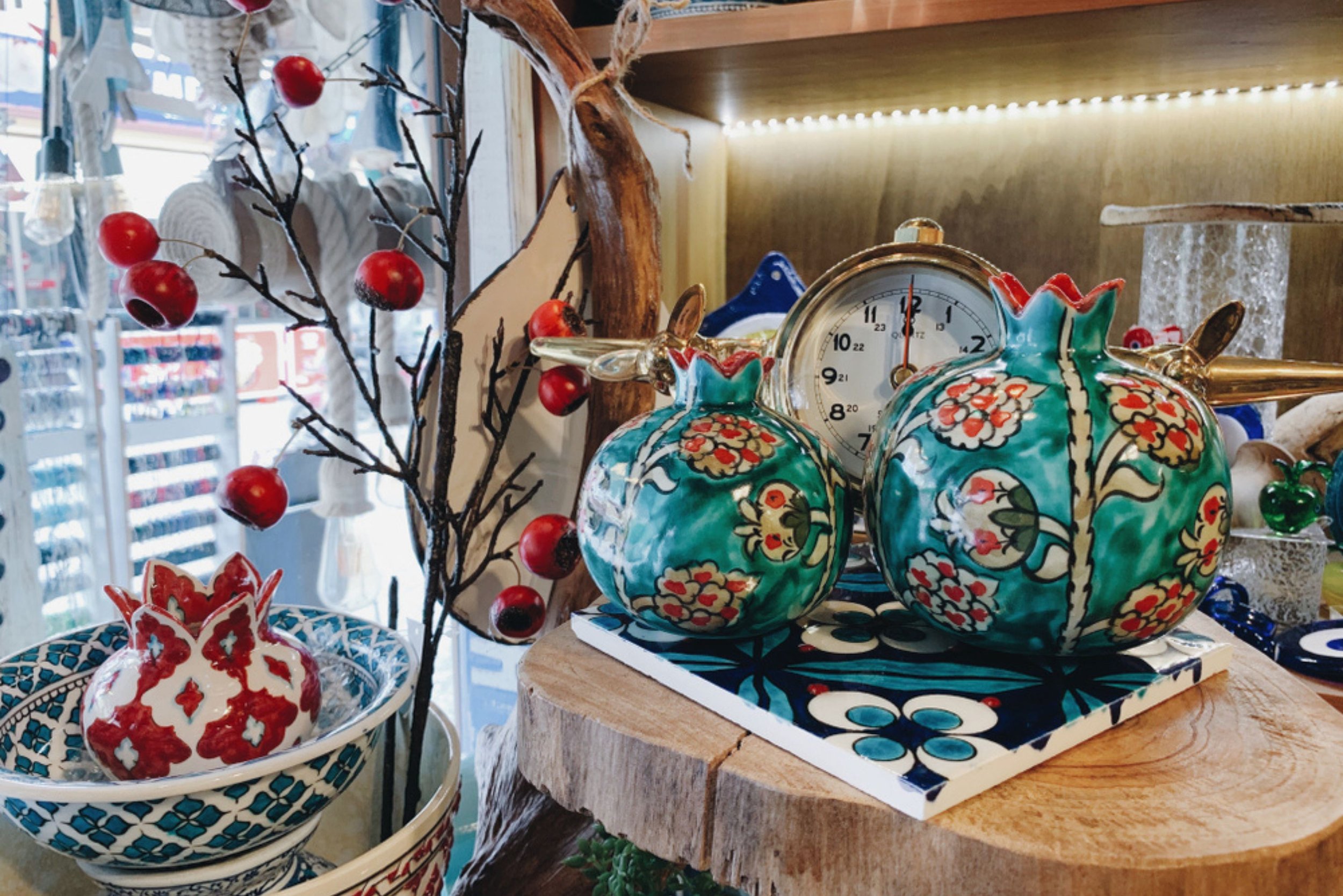 Decorative ceramic pumpkins with floral patterns, a vintage clock, a red and white decorative object in a bowl, and a branch with red berries, displayed on a wooden surface with a patterned mat and a wooden log background.