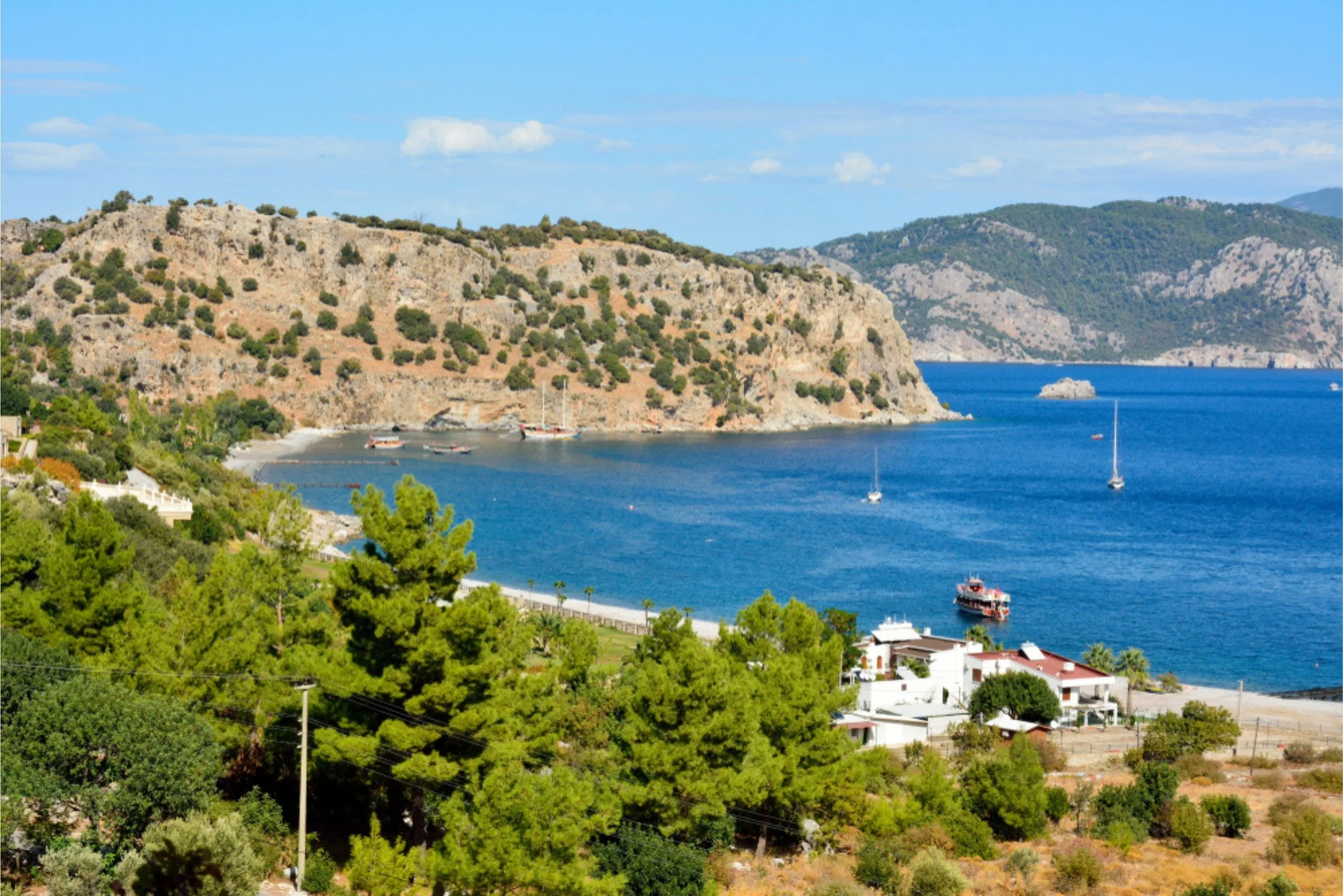 Scenic view of a coastal area with a large hill covered in sparse vegetation, a calm blue bay with boats anchored, and white buildings among green trees in the foreground.