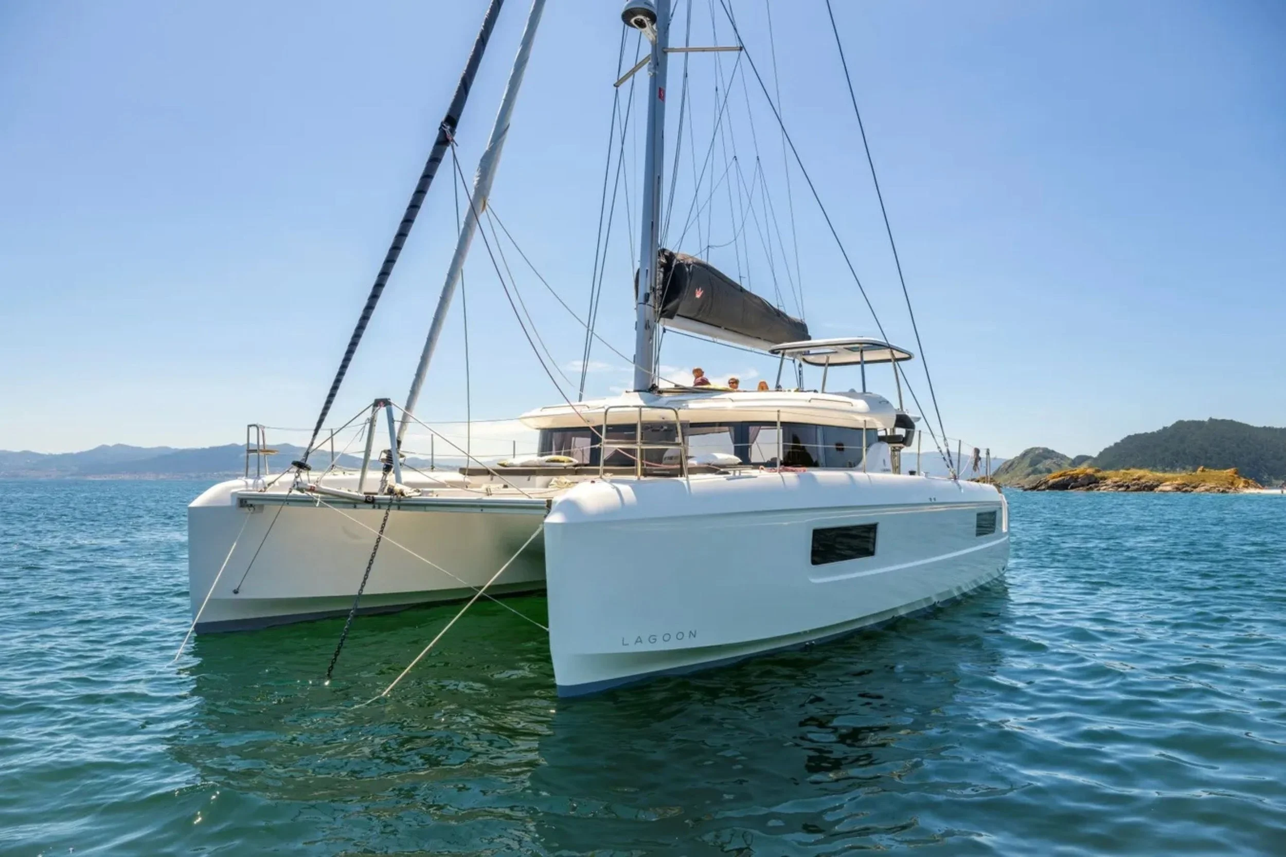 A white sailing catamaran named Lagoon floating on blue water with a clear sky and a distant island with hills in the background.