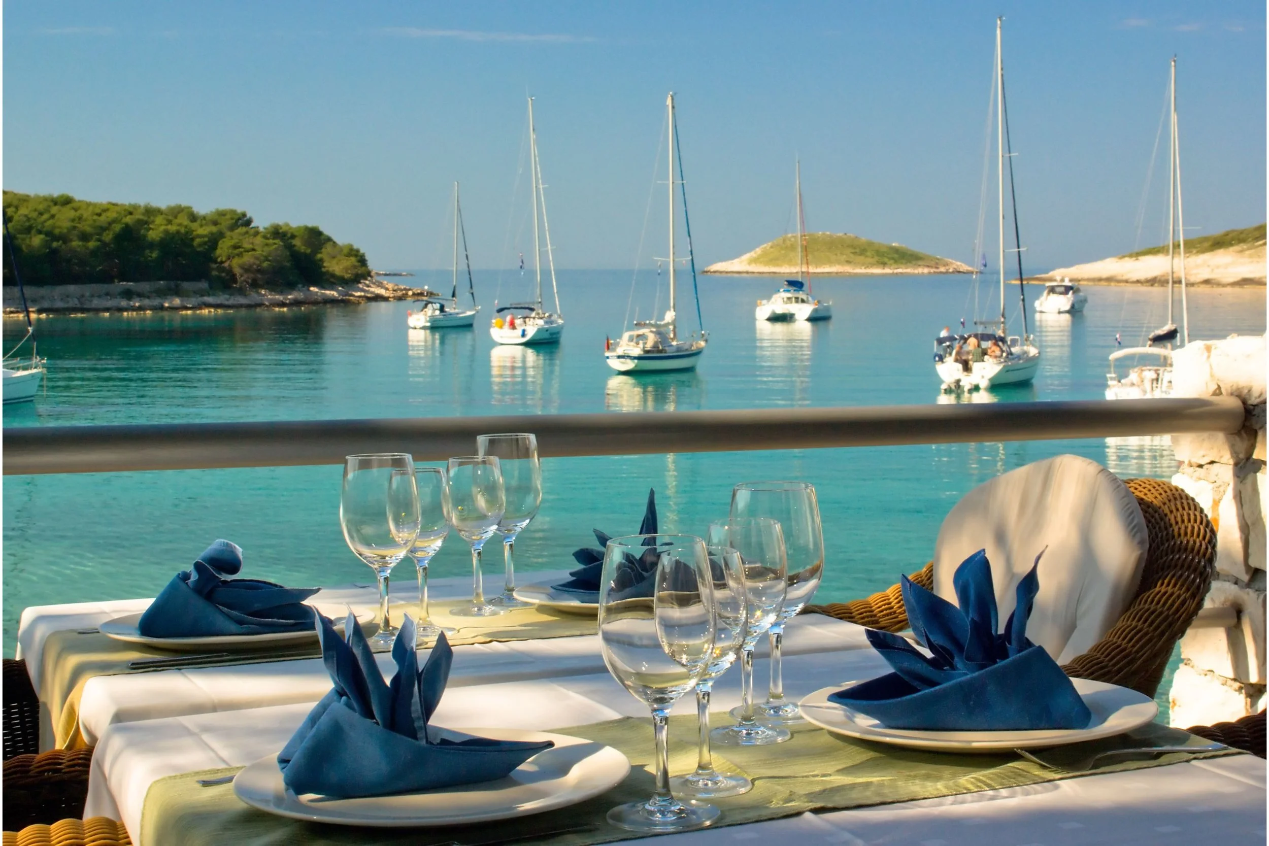 A seaside restaurant table set with wine glasses, blue napkins, and white plates, overlooking a calm harbor with sailboats and a small island in the background.