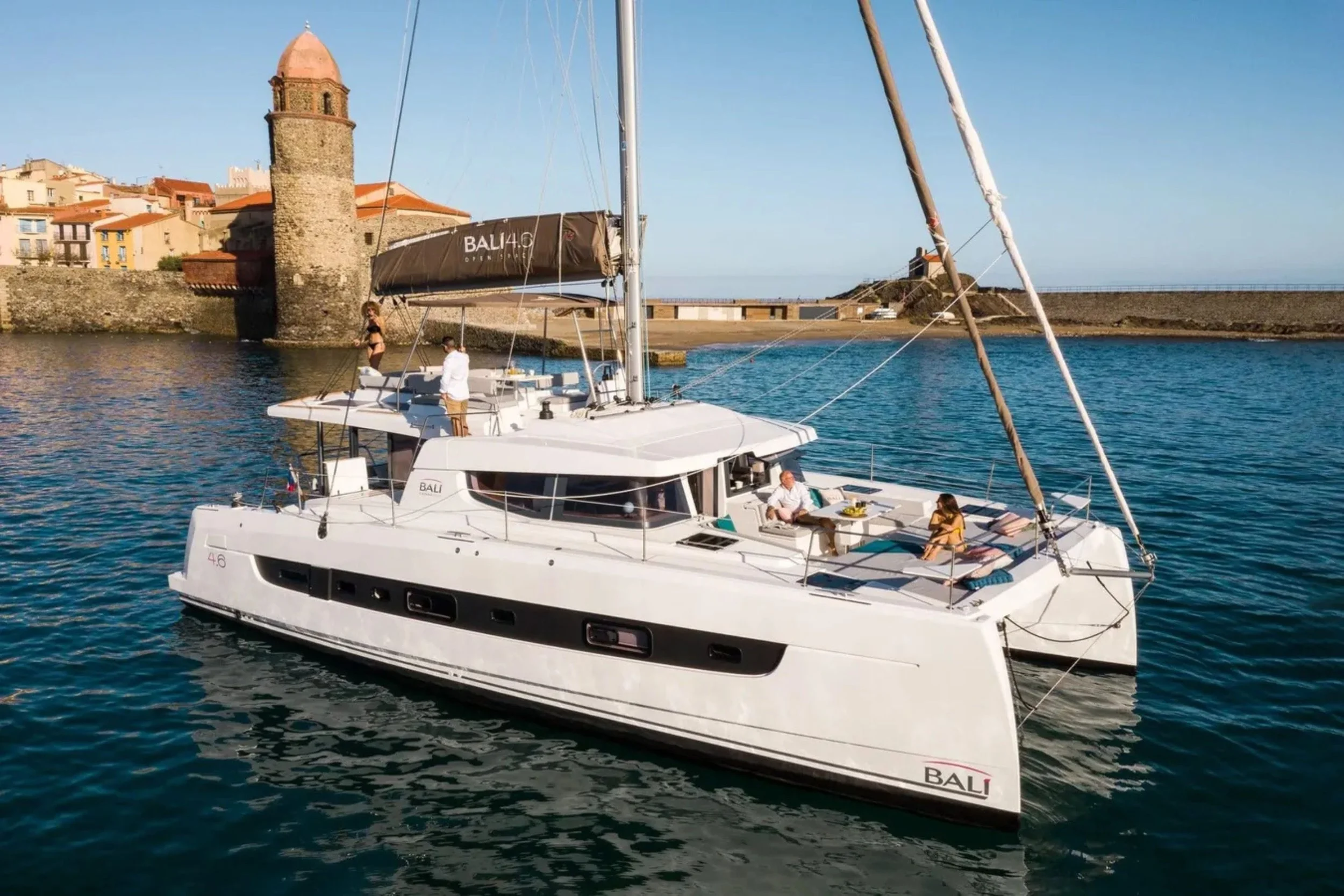 A white sailing yacht named BALI floats on calm blue water near a coastal town with stone buildings and a small tower. Several people are relaxing on the deck, enjoying the sunny weather.
