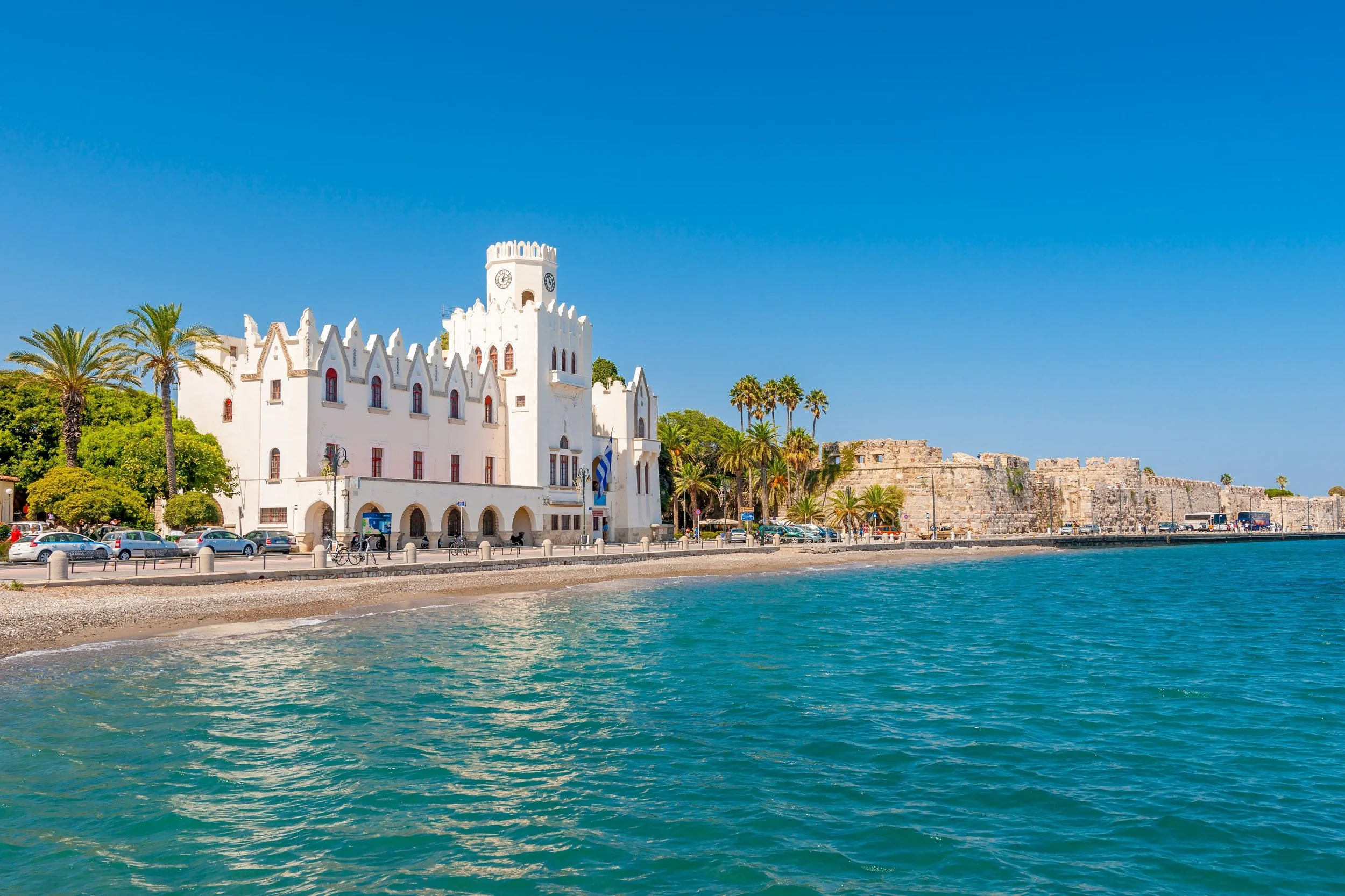 A white castle-like building with a tower, palm trees, parked cars, and a stone wall along a beach with clear blue water and a bright sky.