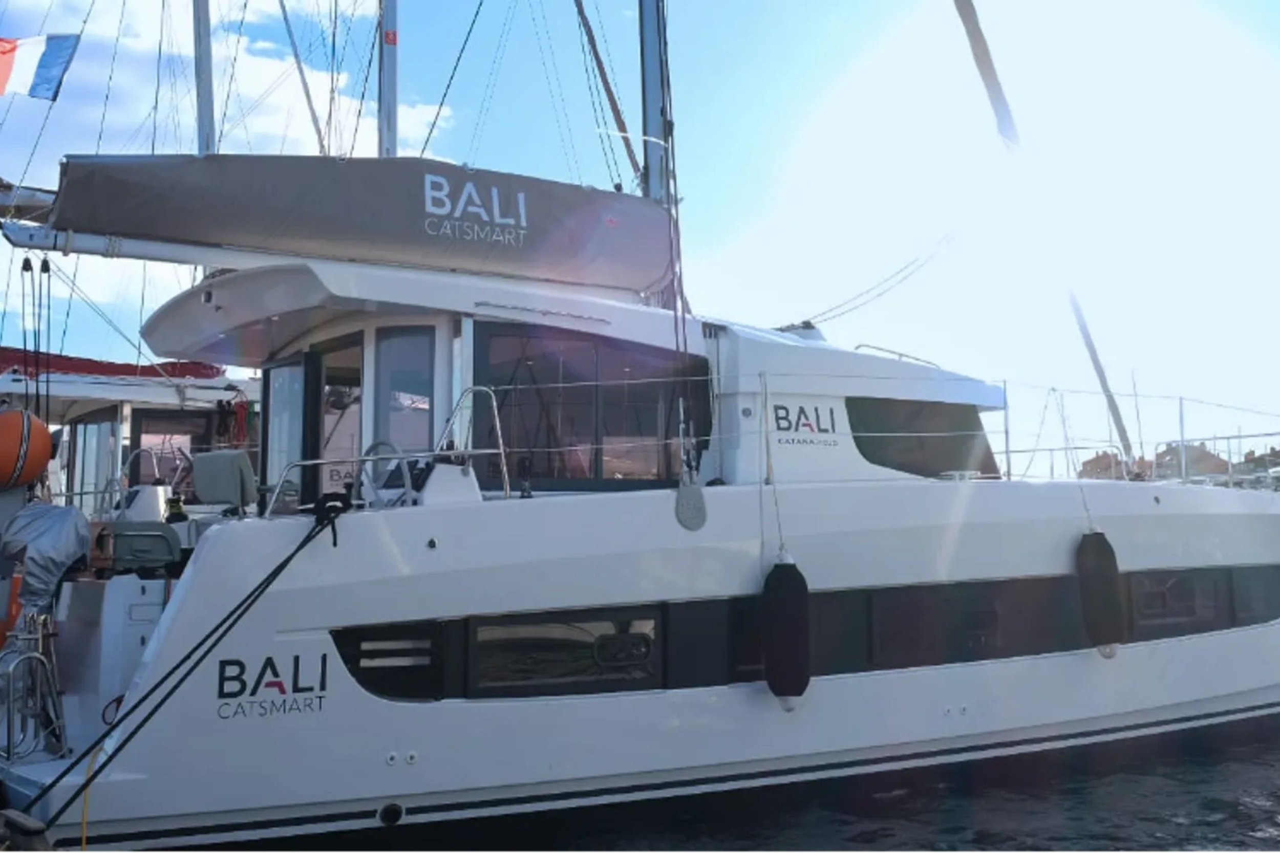 A white sailing yacht docked at a marina with the words 'BALI CATSMART' on the side and on the sail cover.
