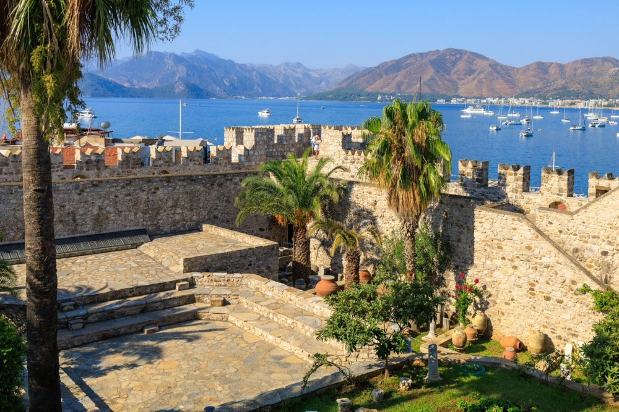 View of a historic stone fortress with stairs leading to a courtyard, palm trees, and potted plants. In the background, a blue lake with sailboats and mountainous terrain.