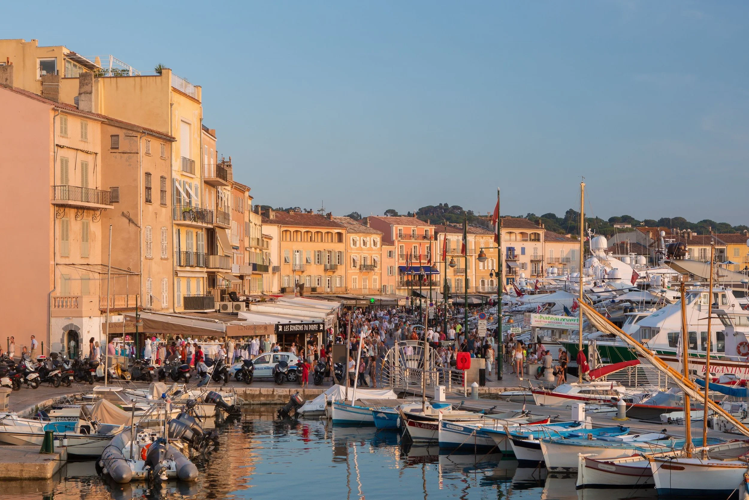 Harbor filled with boats and yachts, promenade with people and shops, pastel-colored buildings, and a clear blue sky.