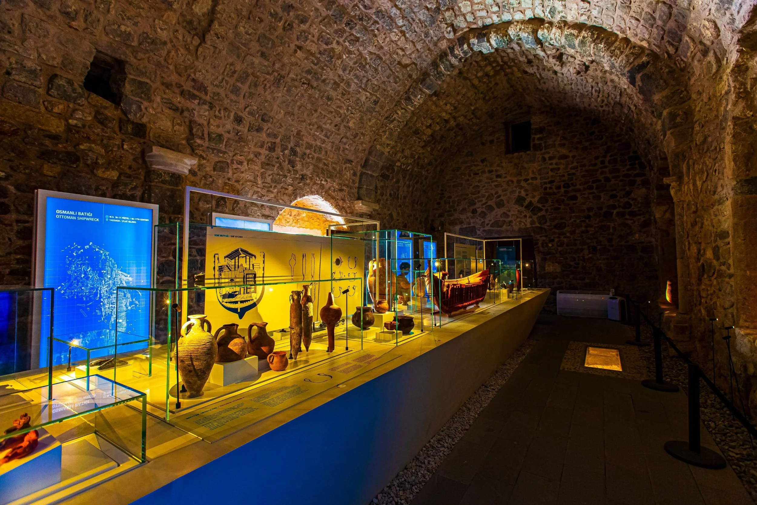 Exhibit of ancient pottery and artifacts inside a stone-walled museum hall with a vaulted ceiling.