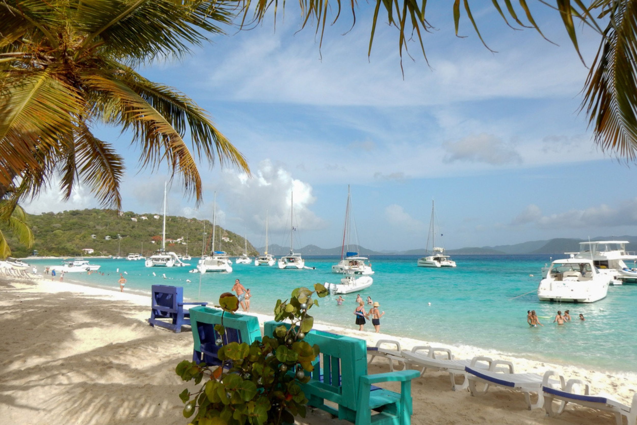 A tropical beach scene with turquoise water, white sand, palm trees, and boats anchored offshore. People are swimming and relaxing by the shore, with colorful benches in the foreground.
