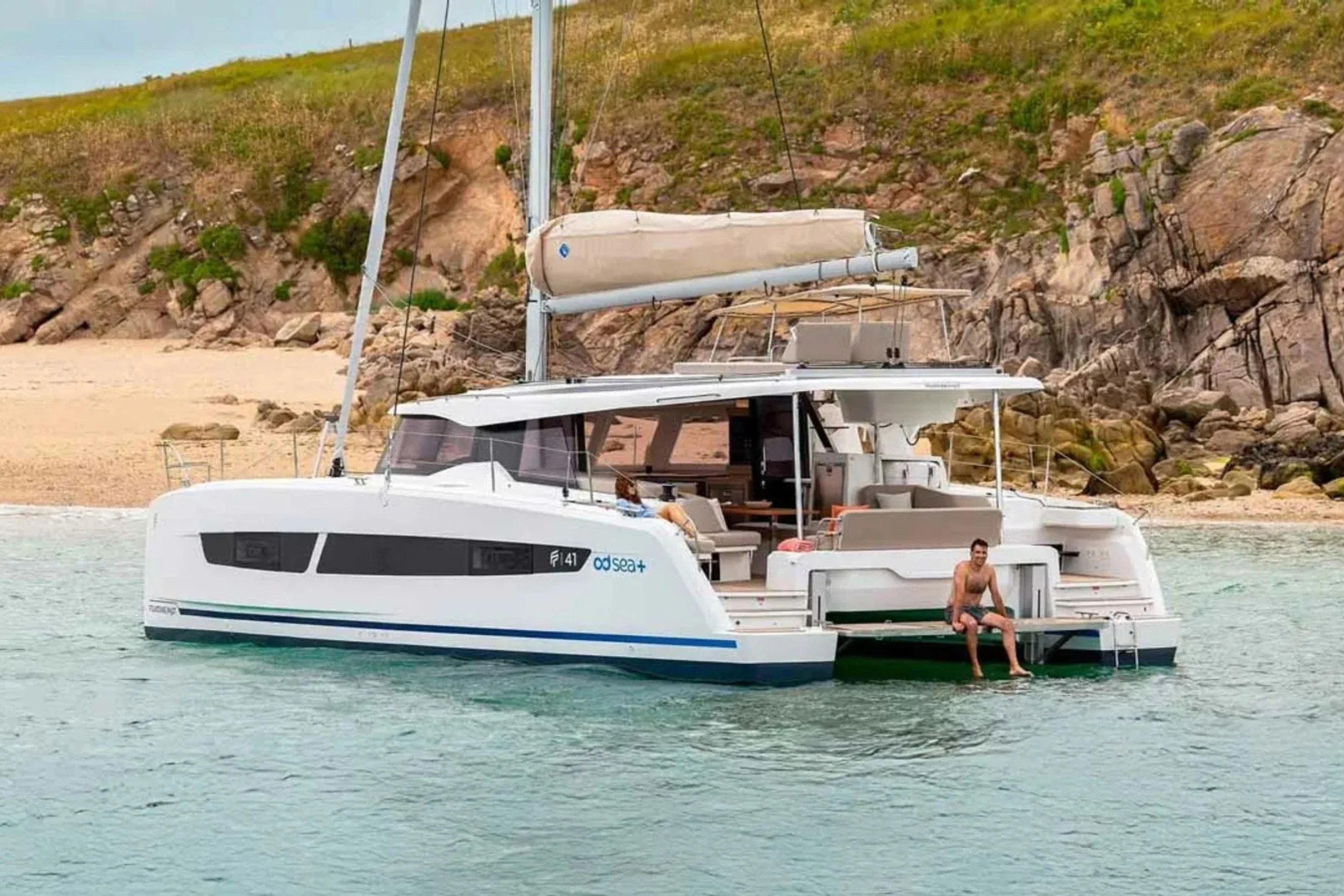 A white modern sailboat anchored near a sandy beach with rocky cliffs in the background. A man is sitting on the boat's swimming platform, with his legs dangling into the water, smiling.