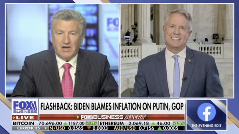 A split-screen television news broadcast featuring two men. The man on the left is a news anchor in a dark suit with a pink tie, and the man on the right is a guest in a blue suit with a light blue tie, standing in front of classical columns and architecture.