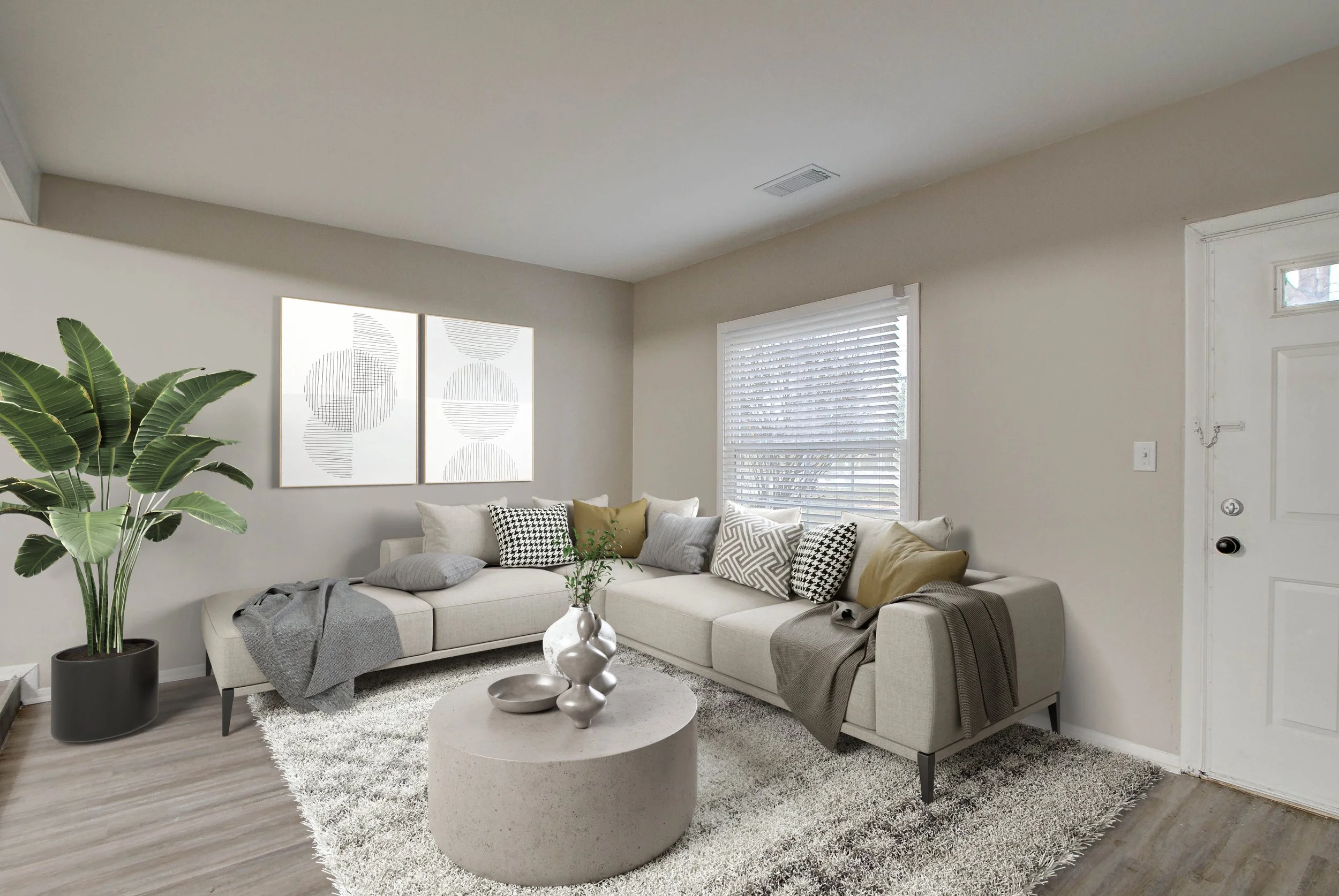 Modern living room with a beige L-shaped sofa, patterned throw pillows, a large potted plant, a white textured rug, a round coffee table with decorative vases, and minimalist wall art. The room has a window with blinds and a white door.