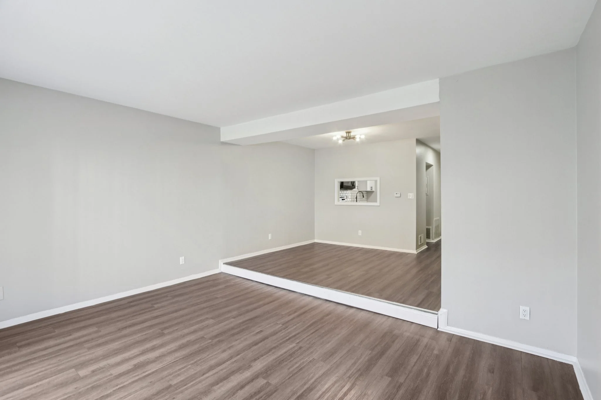 Empty living room with light gray walls, wood laminate flooring, and a small opening to kitchen area.