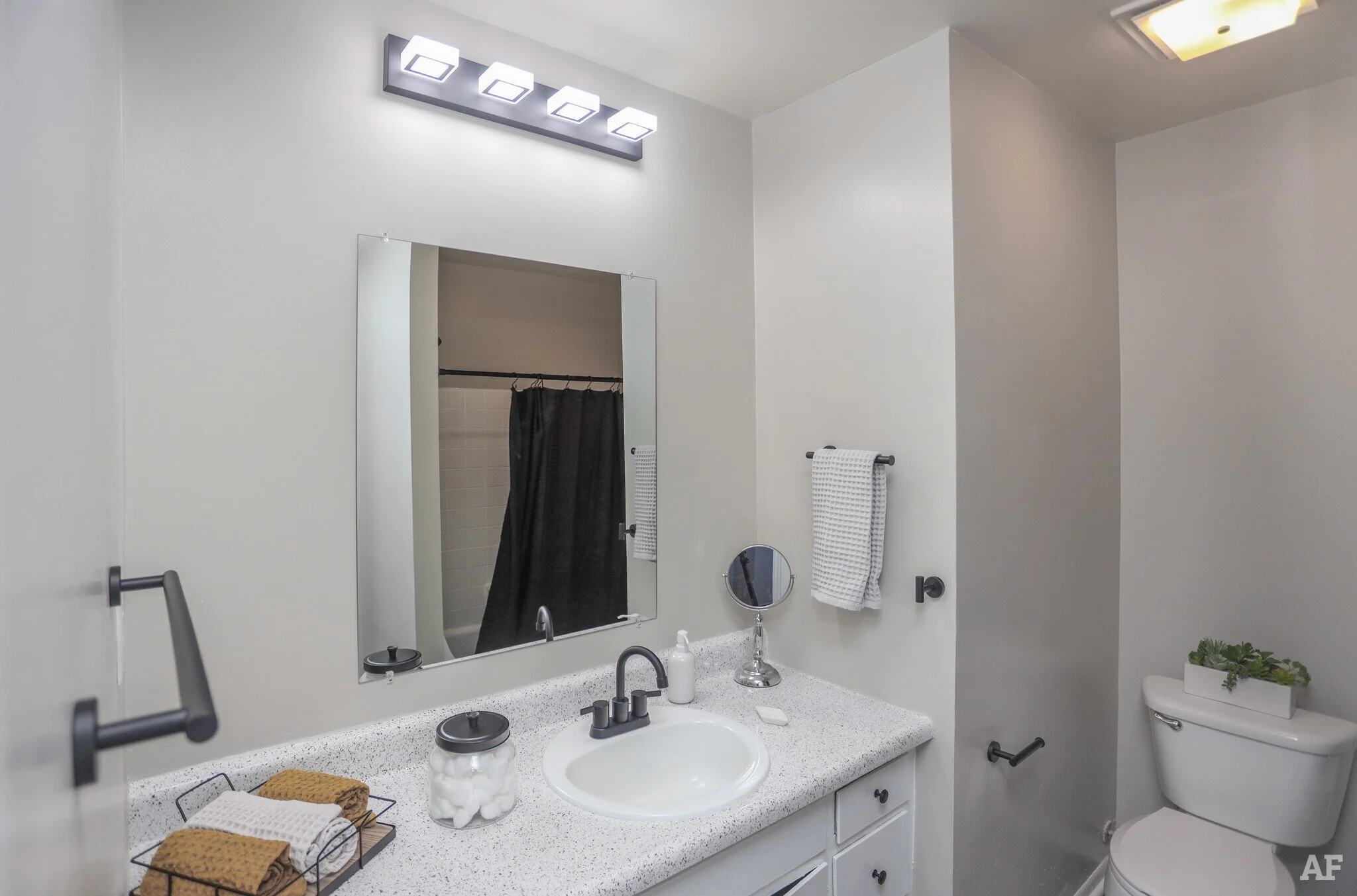 A modern bathroom with a white countertop, a sink with a black faucet, a large mirror, a small round mirror, a soap dispenser, and decorative towels. The toilet has a plant on top. A black towel hangs on a rack, and a black shower curtain is visible