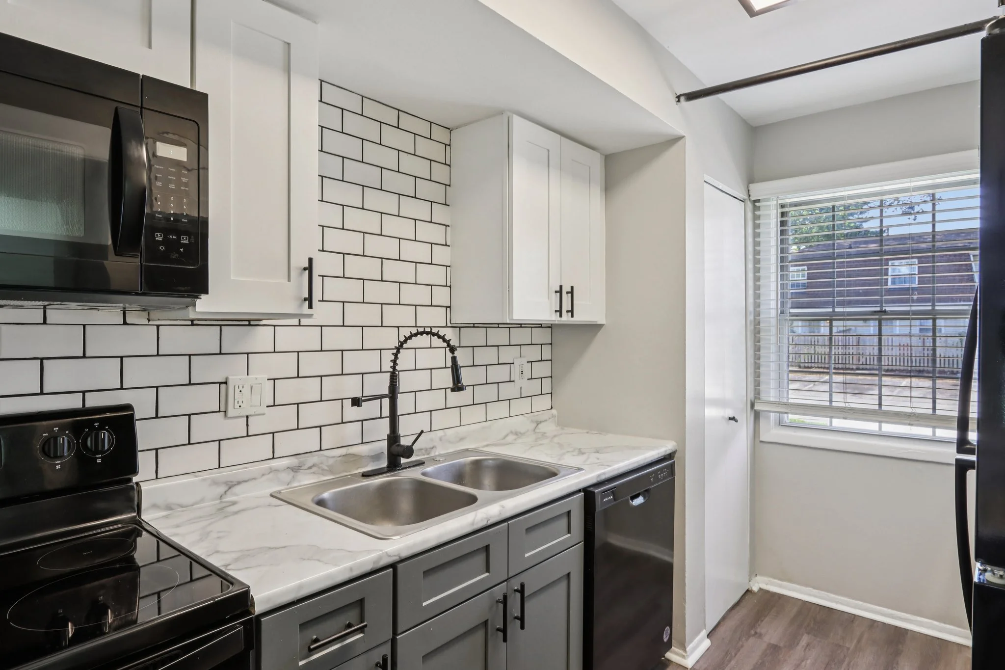 A modern kitchen with a black microwave, black stove, and a black dishwasher. The kitchen has white upper cabinets, gray lower cabinets, a marble countertop, and a white subway tile backsplash. There is a window with blinds letting in natural light.