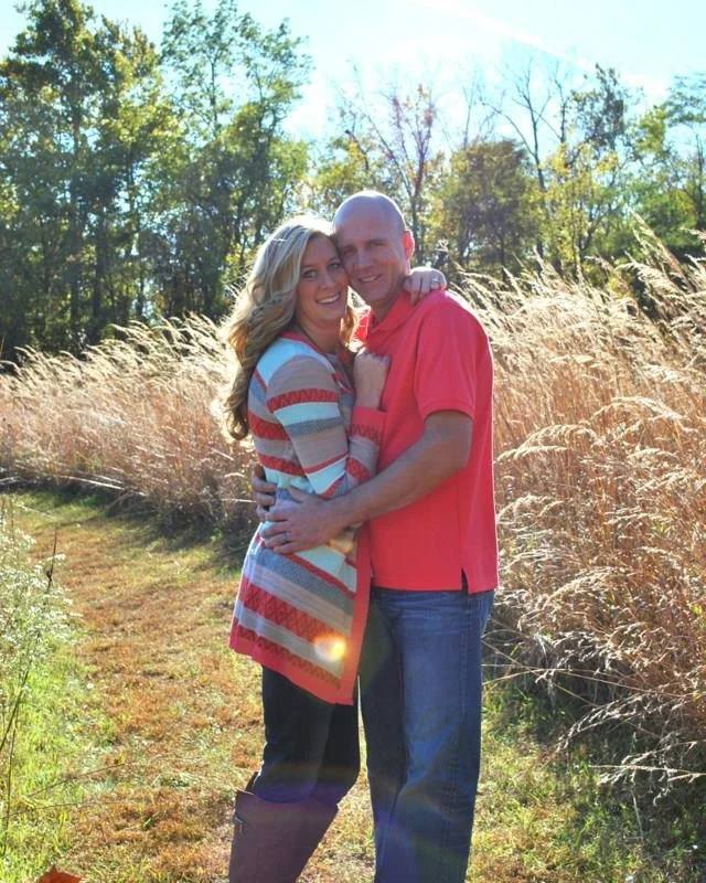 A smiling couple hugging outdoors in a grassy area with trees and tall grass in the background on a sunny day.