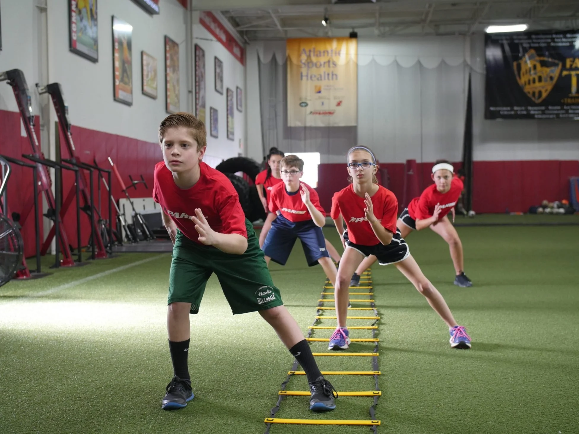 Group of children in athletic clothes practicing agility drills with a ladder on a turf indoors.