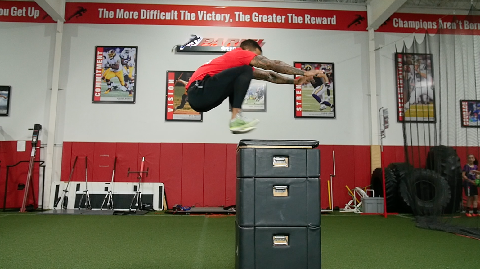 A man in workout attire jumps over a padded box during cross-training in a gym with motivational posters and sports equipment.