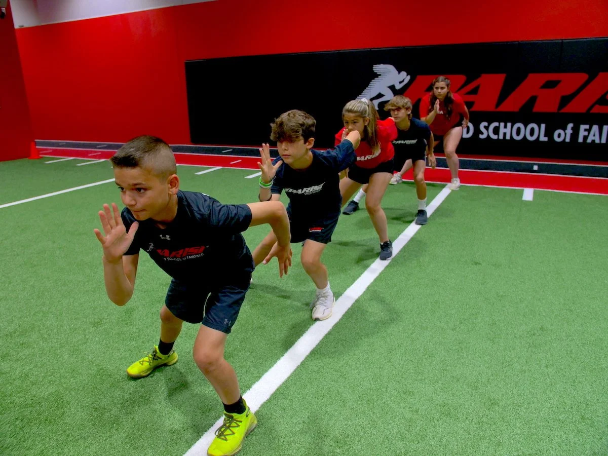 Group of five children in athletic clothing lined up in starting positions on indoor turf field, preparing for a race.