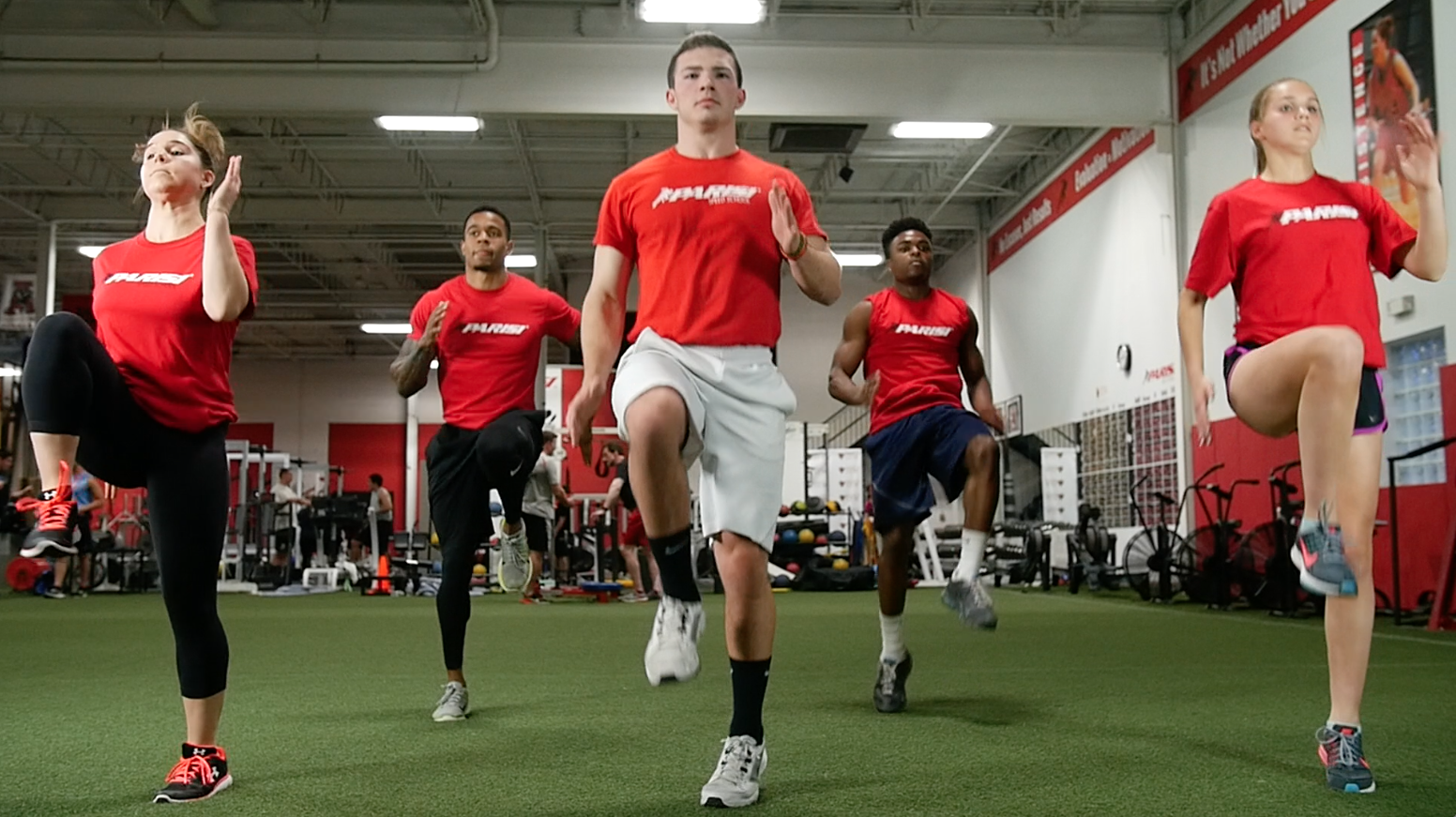 Group of five young athletes in red shirts and activewear practicing balance exercises on one leg in a gym.