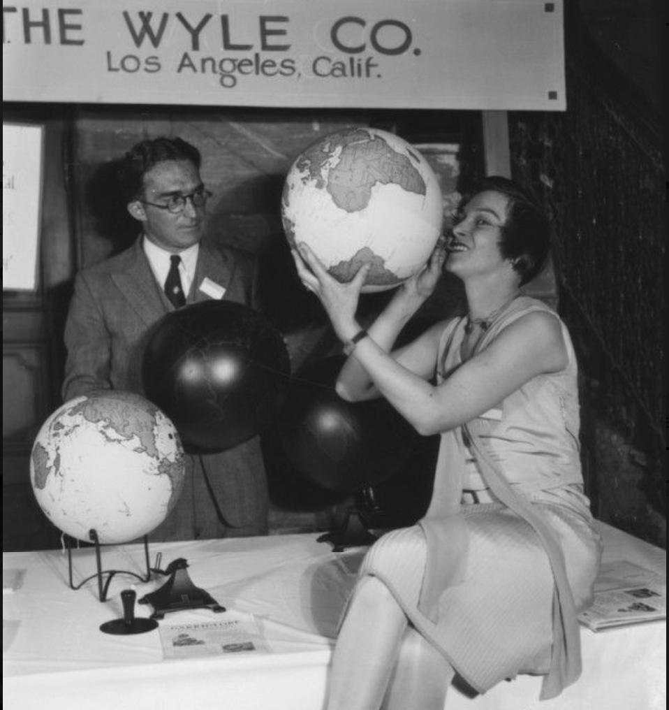 A woman playfully holds a globe near her face while seated at a table with two globes and balloons displaying maps. A man in glasses and a suit stands nearby, observing. The setting appears to be an exhibition or event related to the Wyle Company in Los Angeles, California.