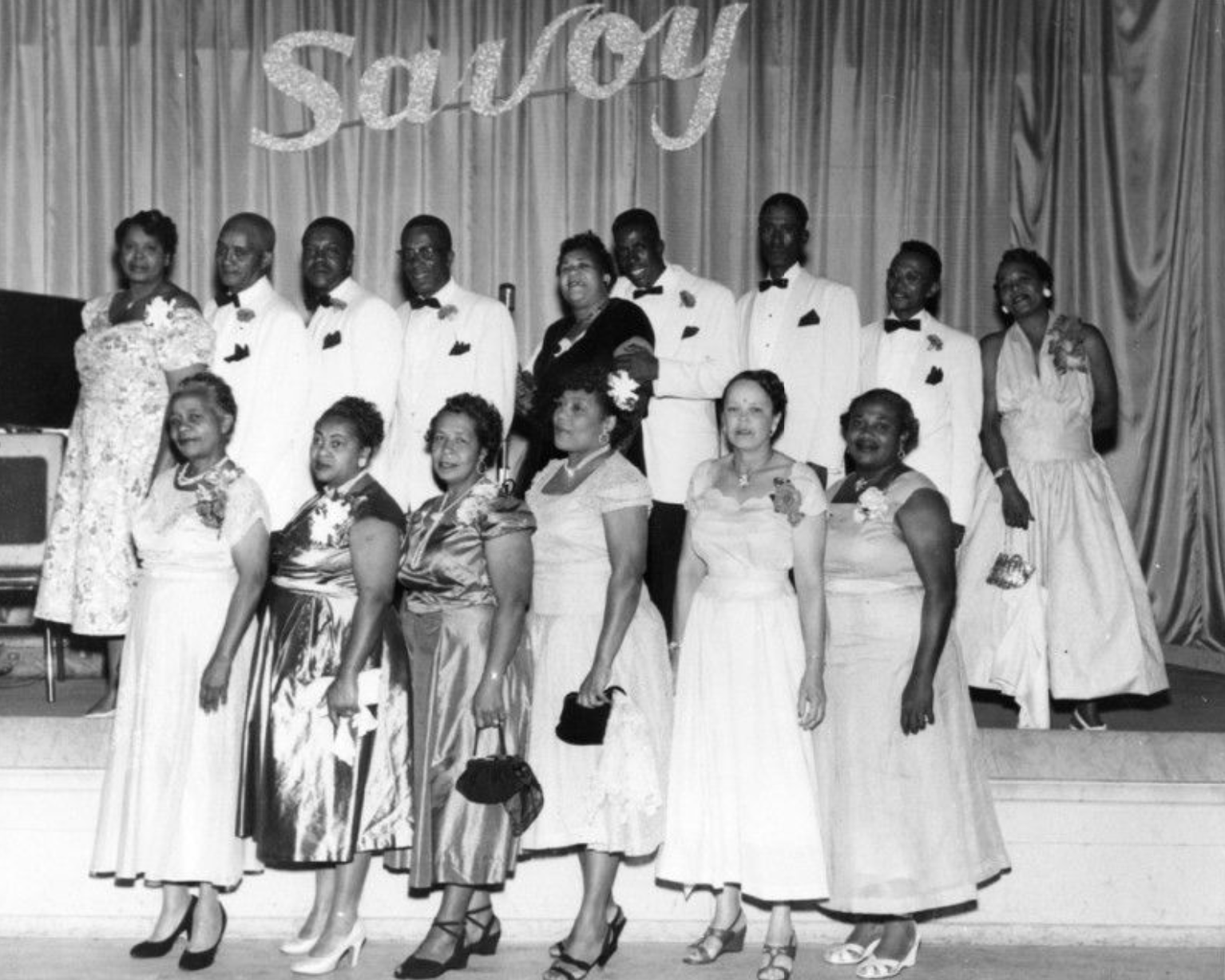 An archival photo of a group of Black adults in formal attire posing for a photo on a stage with a curtain background. The word 'Savoy' is displayed in glittery letters above them.