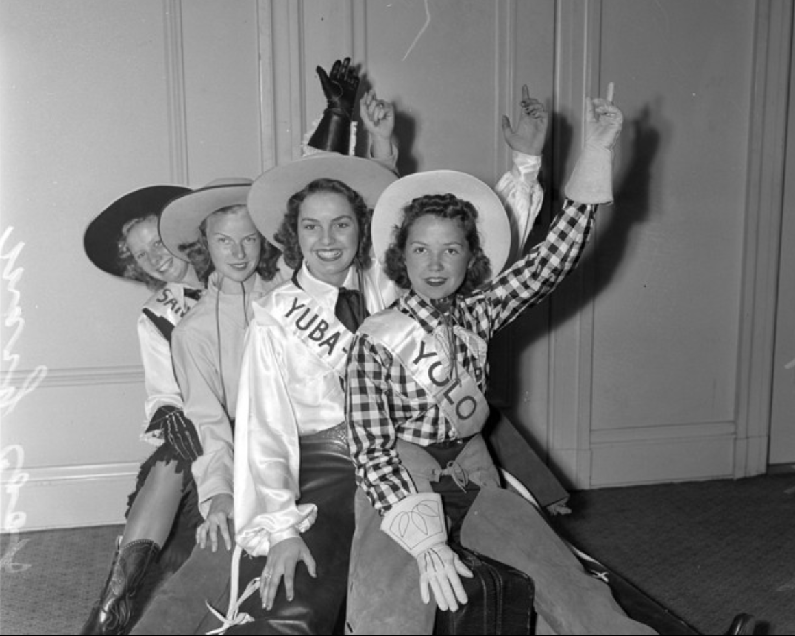 Archival photo of four women dressed in vintage cowgirl attire, wearing sashes with names, and wide-brimmed hats, with some raising their hands, sitting on the floor against a paneled wall.