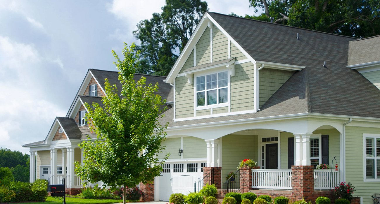 Front view of a two-story, light green house with white trim, a covered porch, and a landscaped lawn with bushes and a tree.