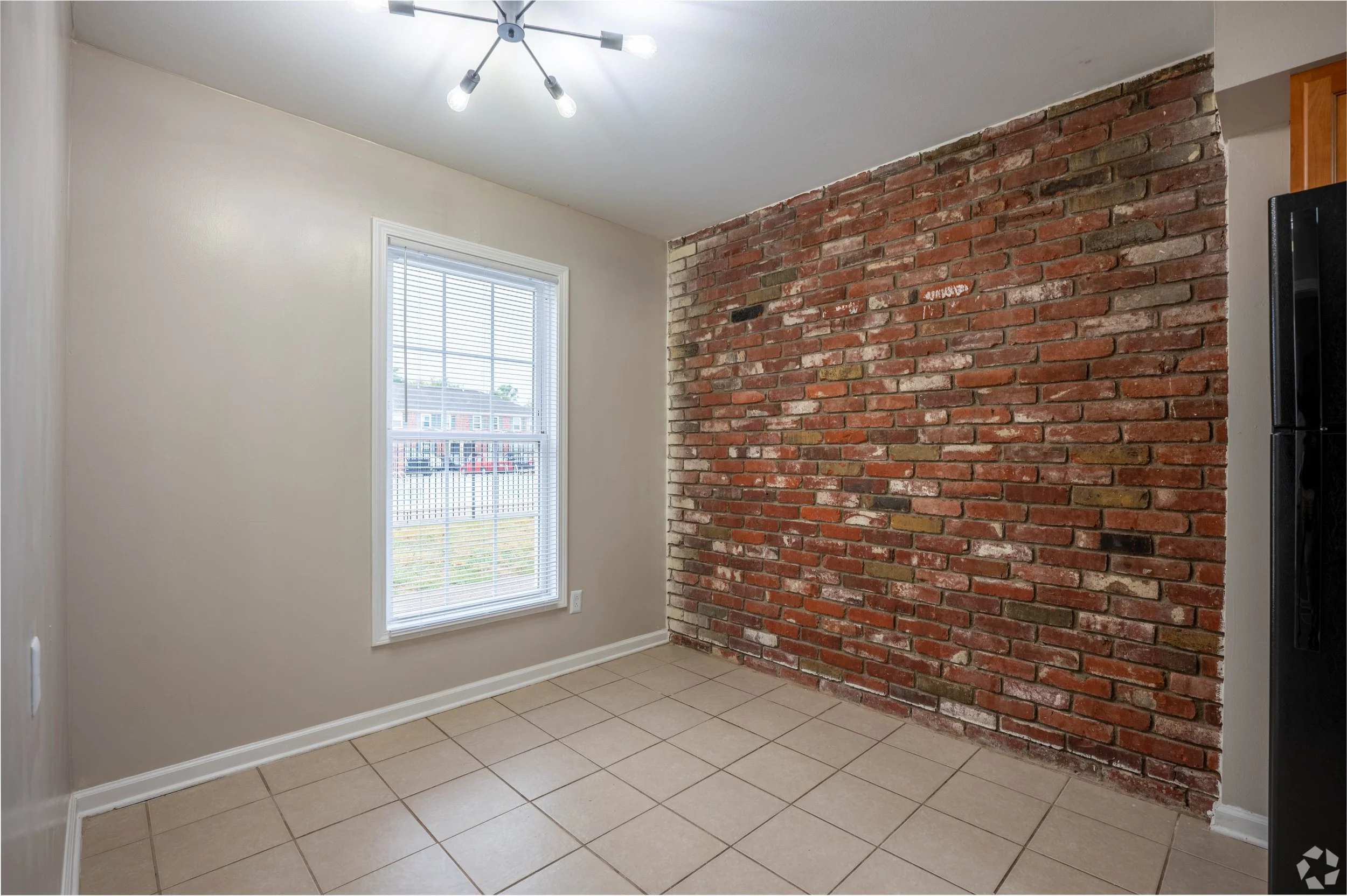 Empty room with beige walls, tiled floor, a window with blinds, a brick accent wall, and ceiling lights.