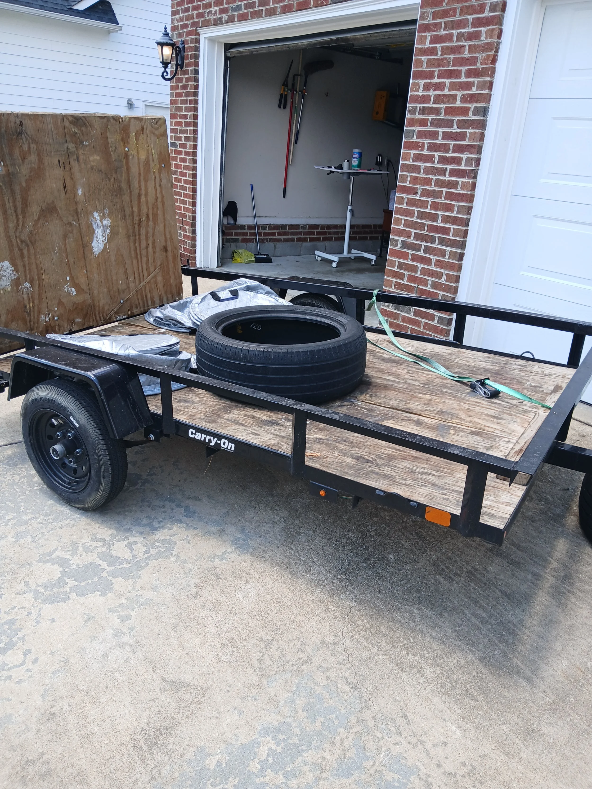 A flatbed trailer with a tire, some tarp, and a green strap is parked outside a garage with tools hanging on the wall.