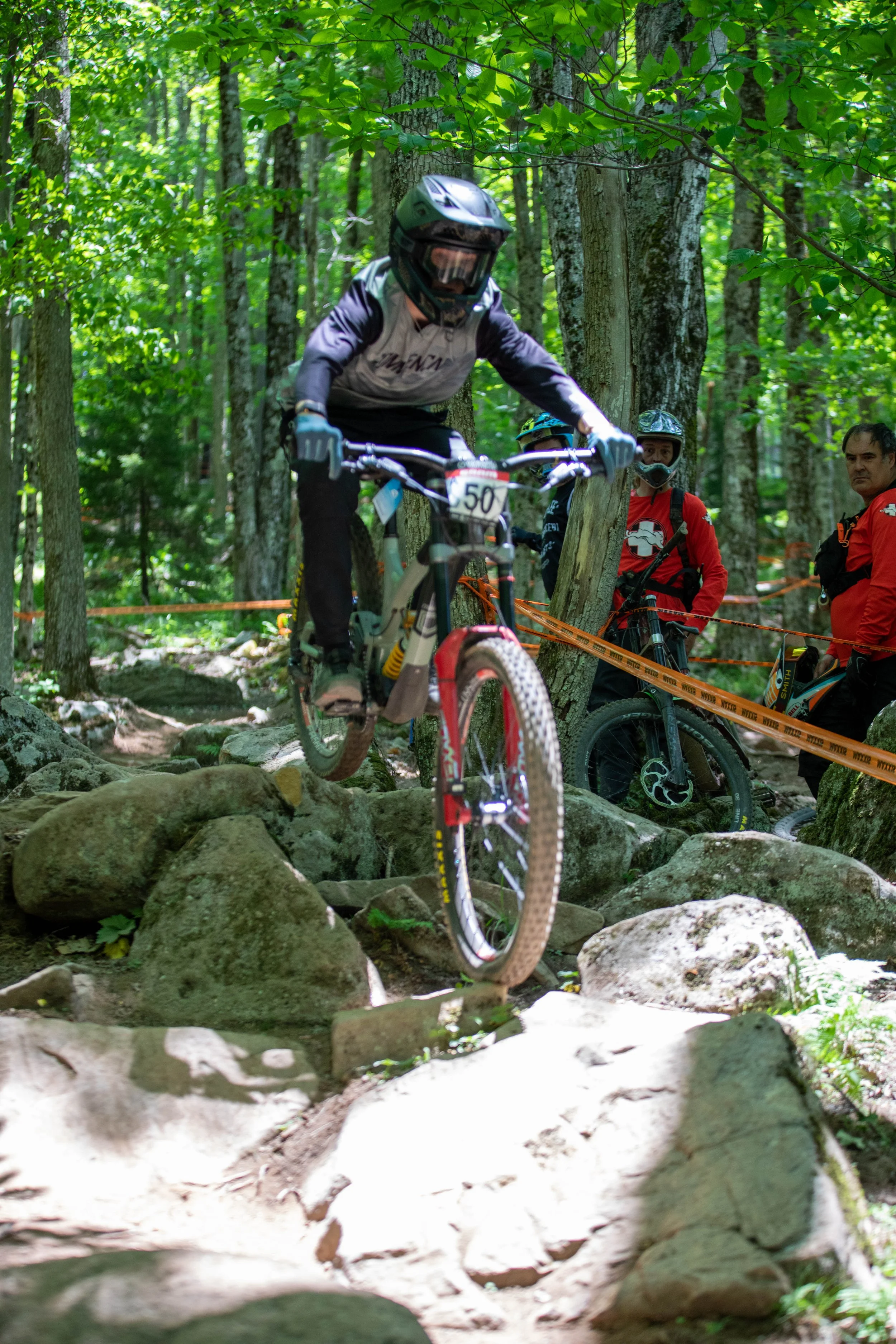 A mountain biker in a black helmet and gloves riding over rocks on a forest trail, with three other cyclists and volunteers watching behind orange safety tape.