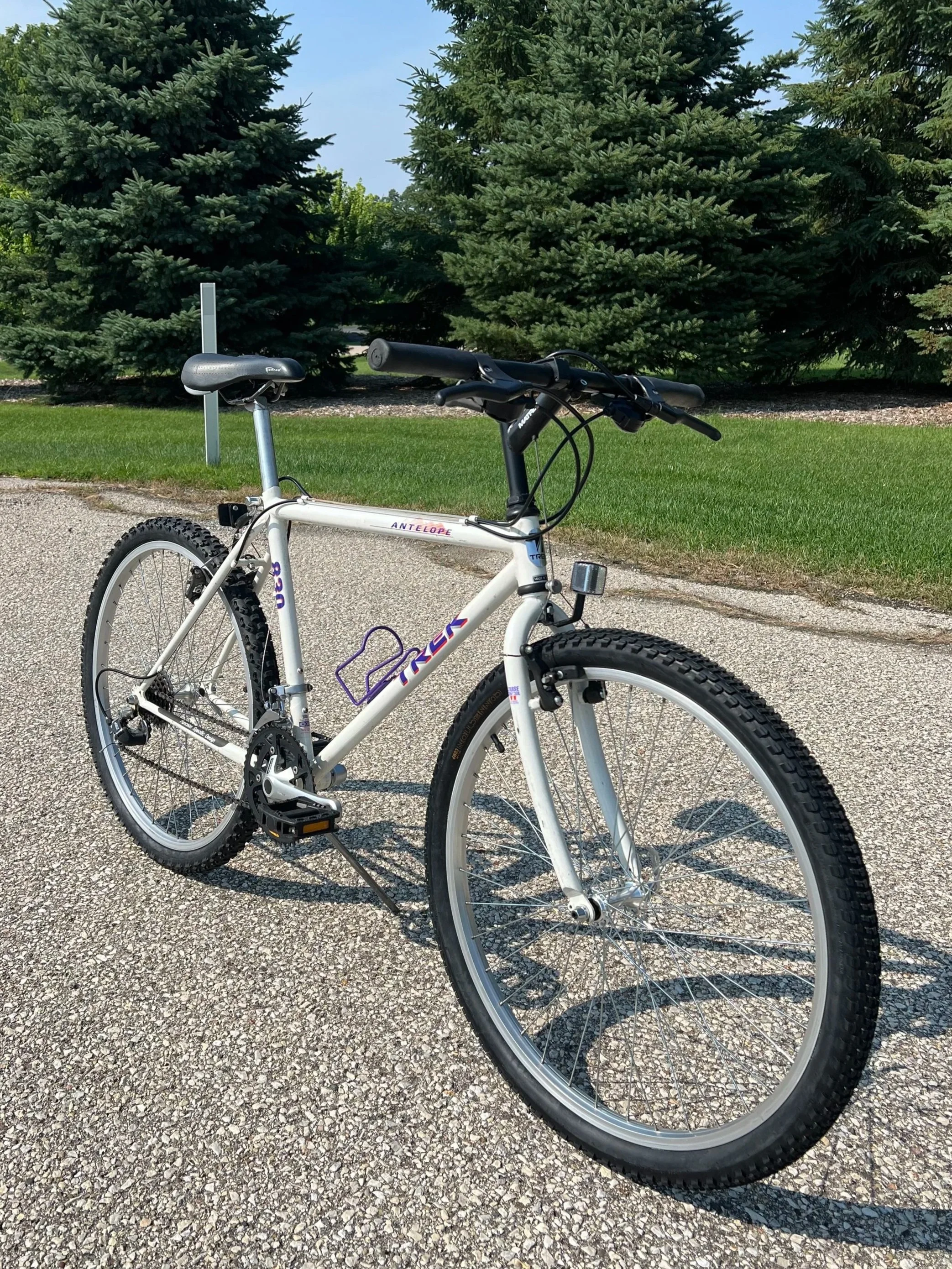 A white Trek Antelope mountain bike parked on a gravel path with evergreen trees in the background.