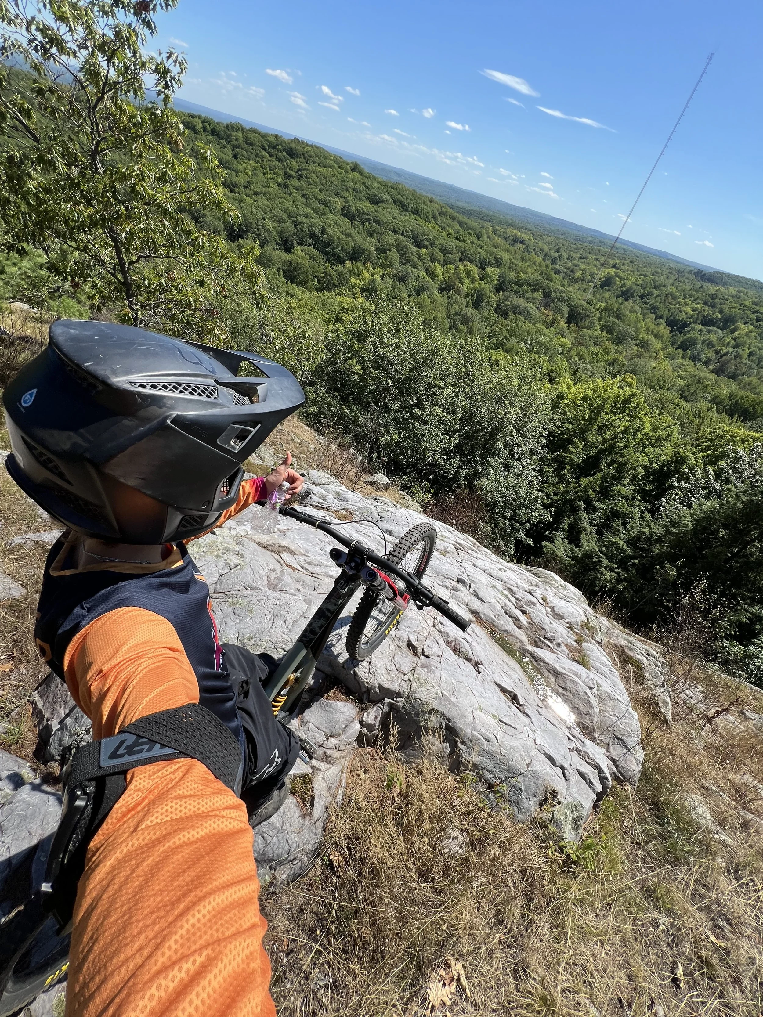 A mountain biker in orange and black gear, wearing a full-face helmet, taking a selfie with a mountain and forested landscape in the background.
