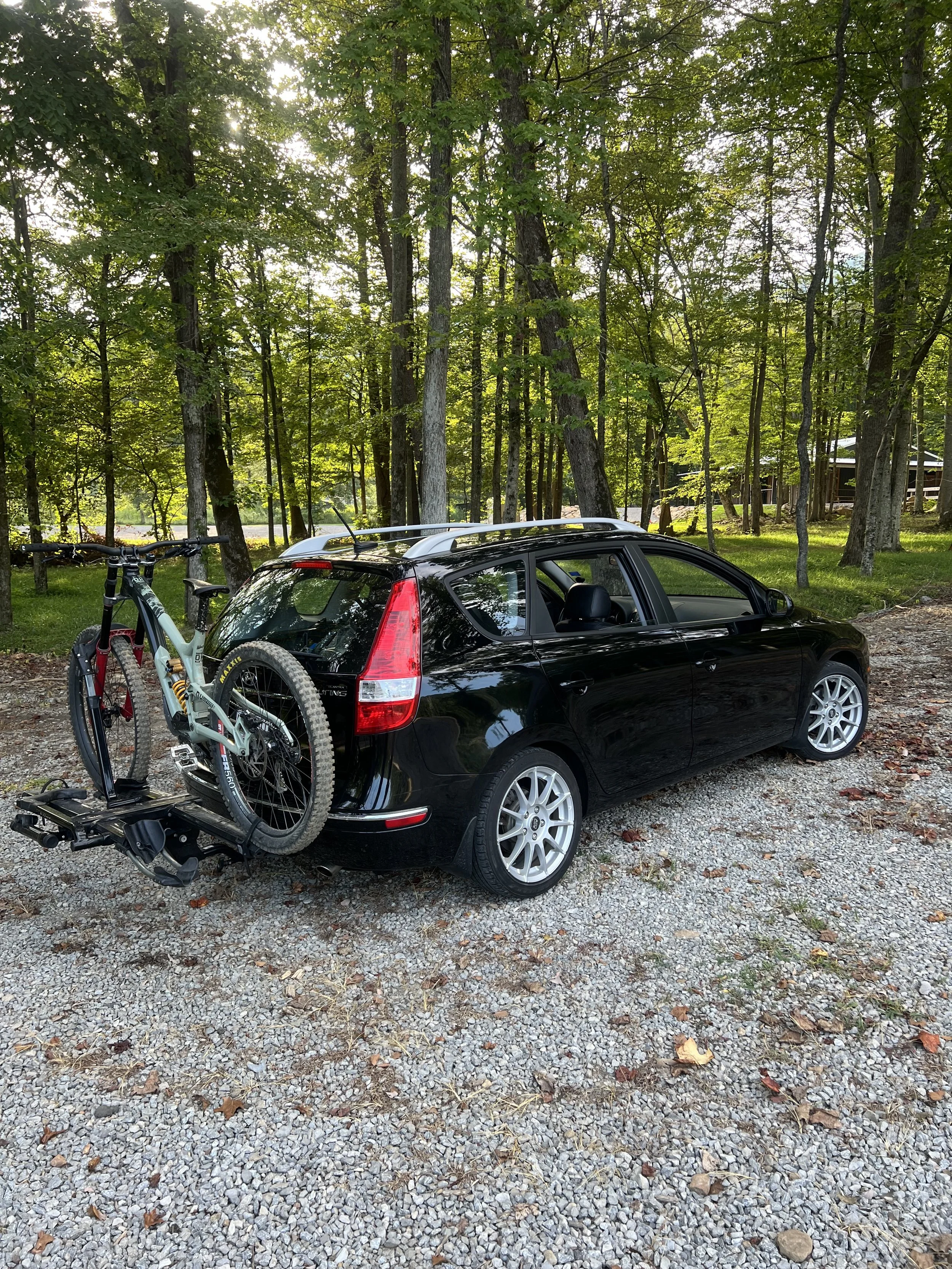 Black station wagon parked on gravel with a mountain bike attached to the rear, surrounded by tall trees in a forested area.