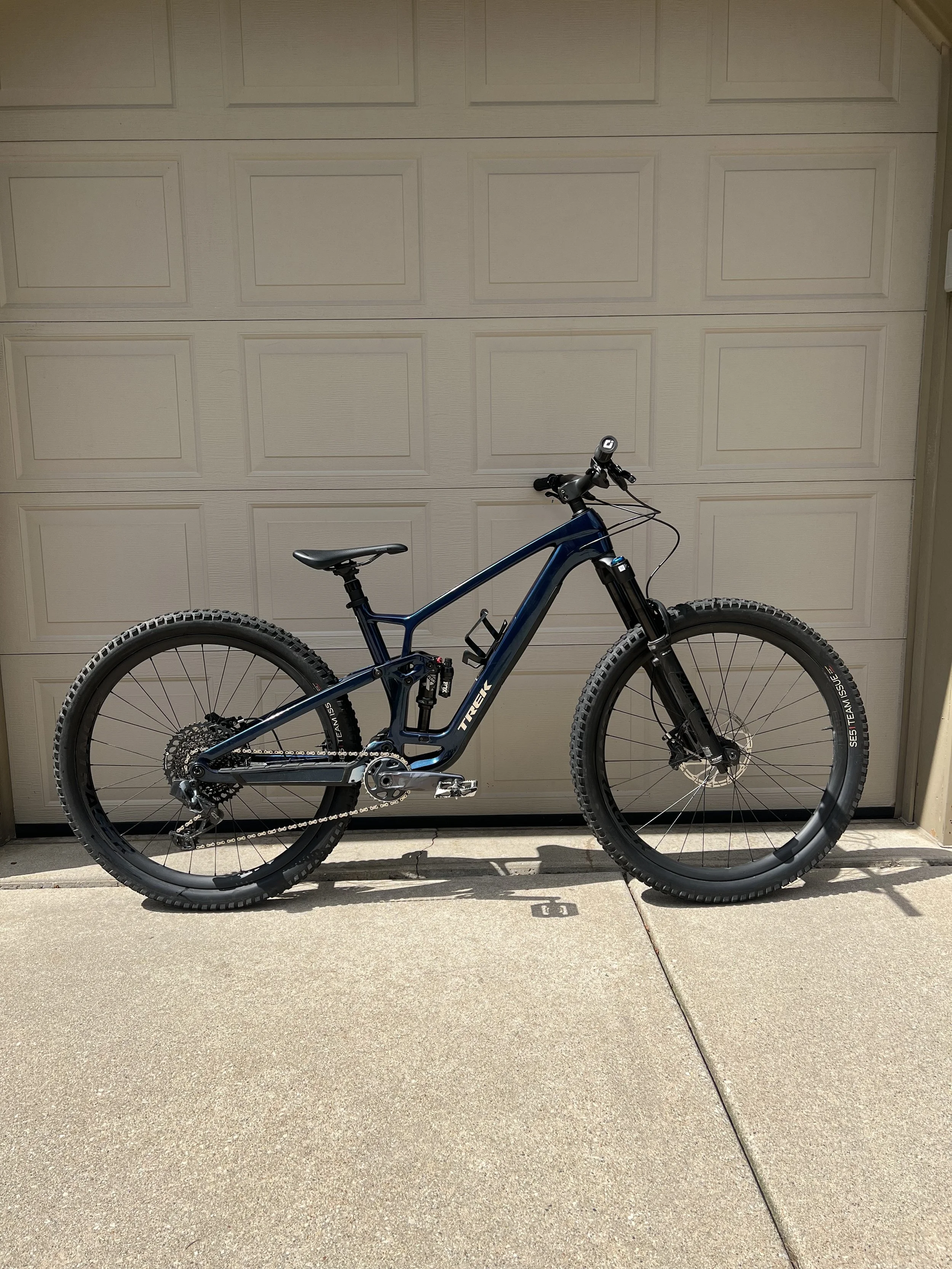 A black and blue Trek mountain bike parked in front of a beige garage door on a concrete driveway.