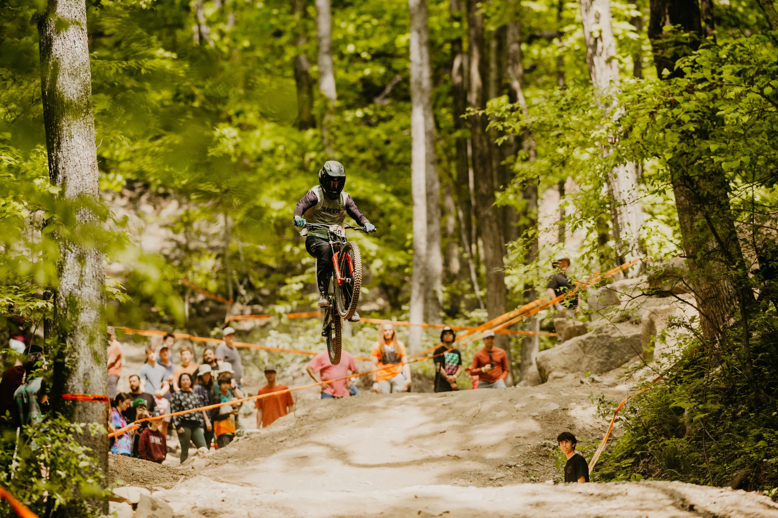 A mountain biker in a black helmet and gear jumps off a dirt ramp during a downhill race in a forest with green leaves, while spectators watch from behind orange safety tape.