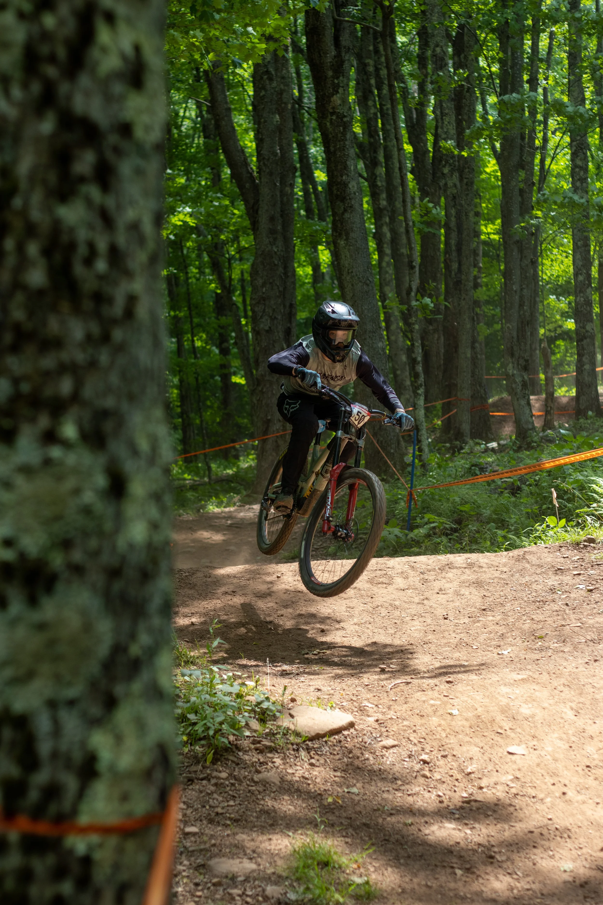 A mountain biker wearing a black helmet and black protective gear jumps off a dirt trail through a forest with lush green trees.
