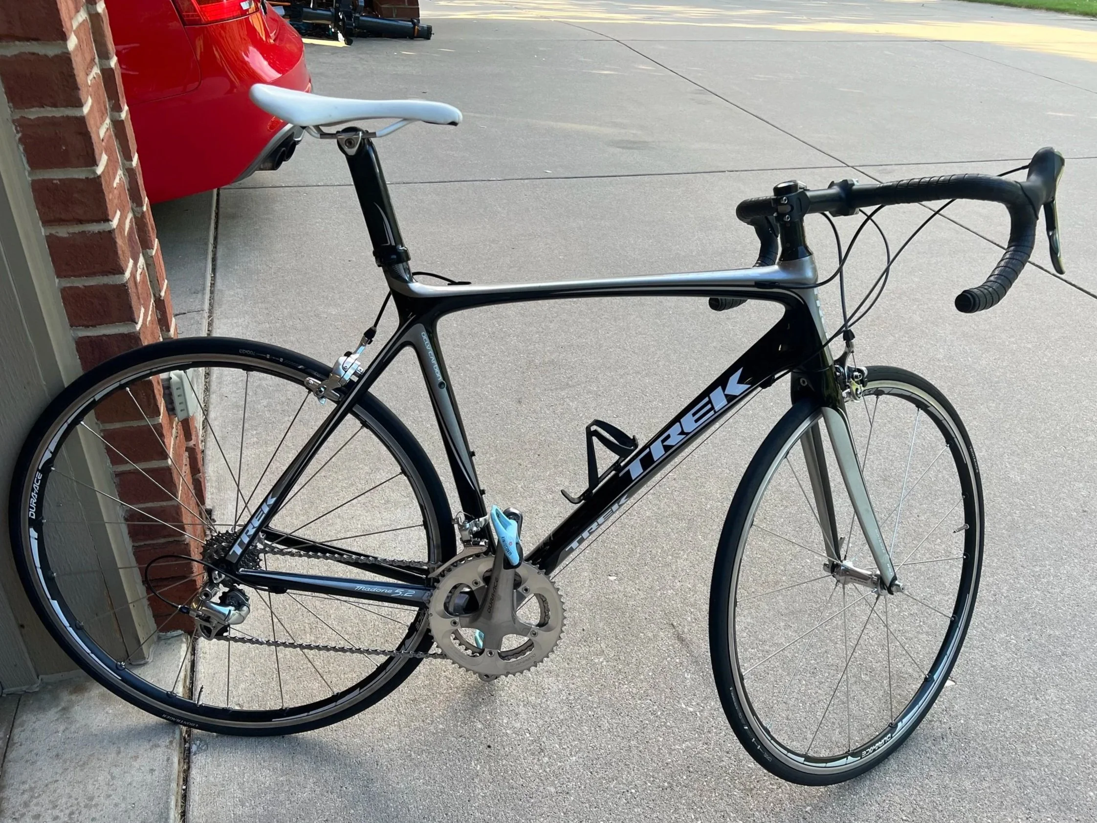 A Trek road bicycle with black and silver frame, drop handlebars, and thin tires, leaning against a brick wall outdoors.