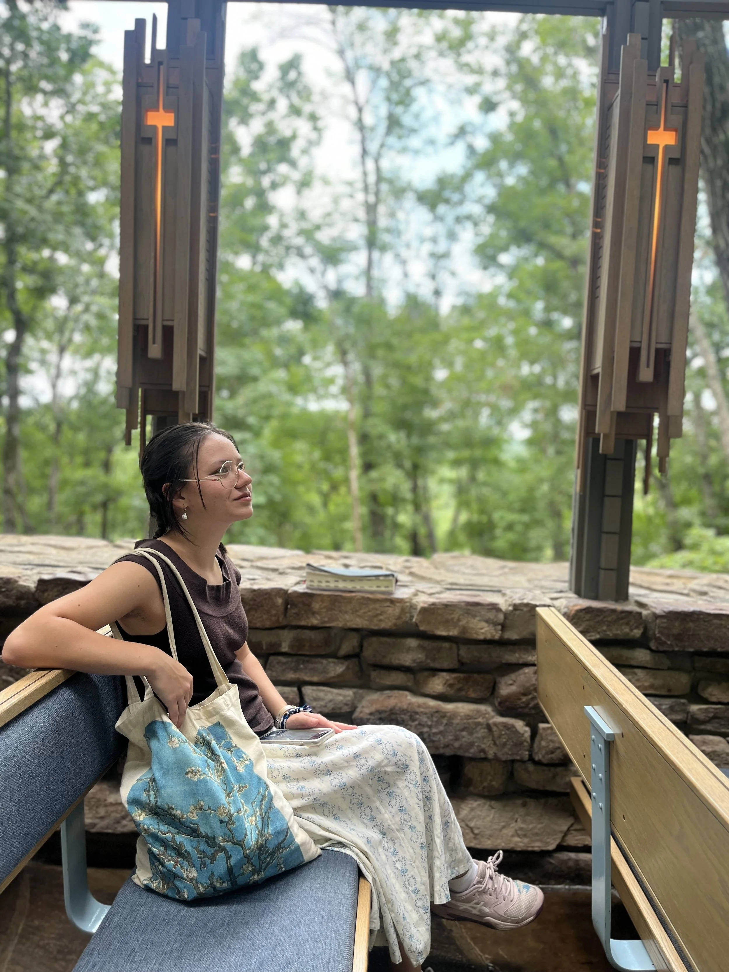 seated on a church bench inside a chapel, with green forest visible through large windows behind her. She is wearing glasses, a dark sleeveless top, a white patterned skirt, and sneakers, holding a tote bag with a blue floral design and a book or notebook on her lap.